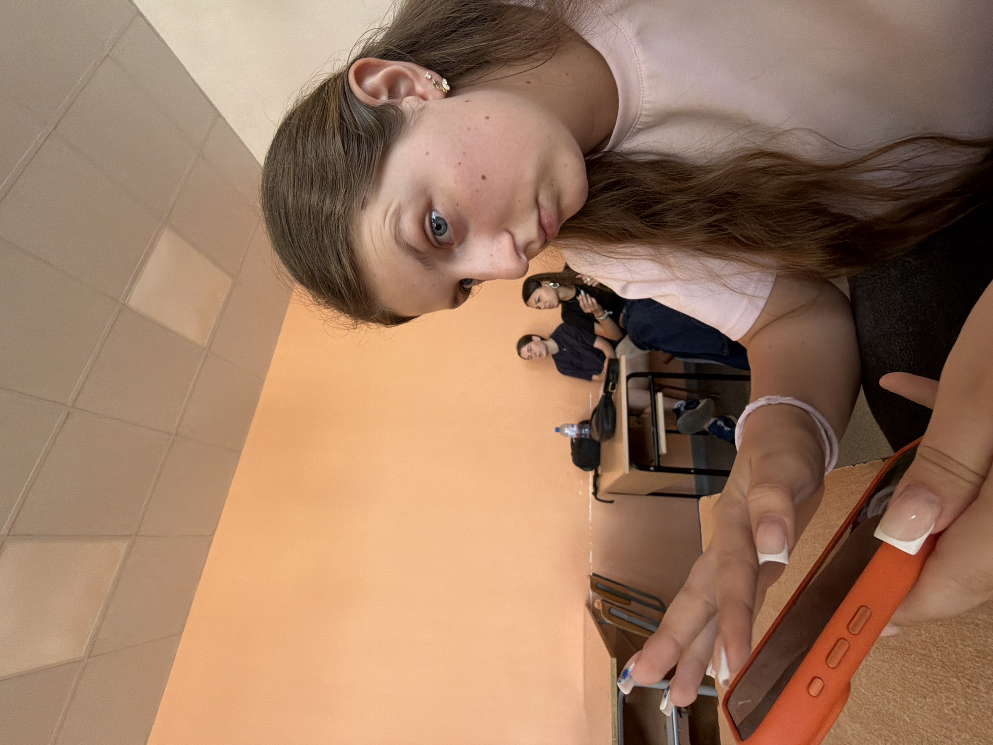 A young woman with long brown hair and blue eyes holds a smartphone in a classroom setting, likely at Burgas Free University in Burgas, Bulgaria. Two other people sit at desks behind her. A bag is visible on the floor by a desk. Walls are painted in a combination of peach and cream colors.