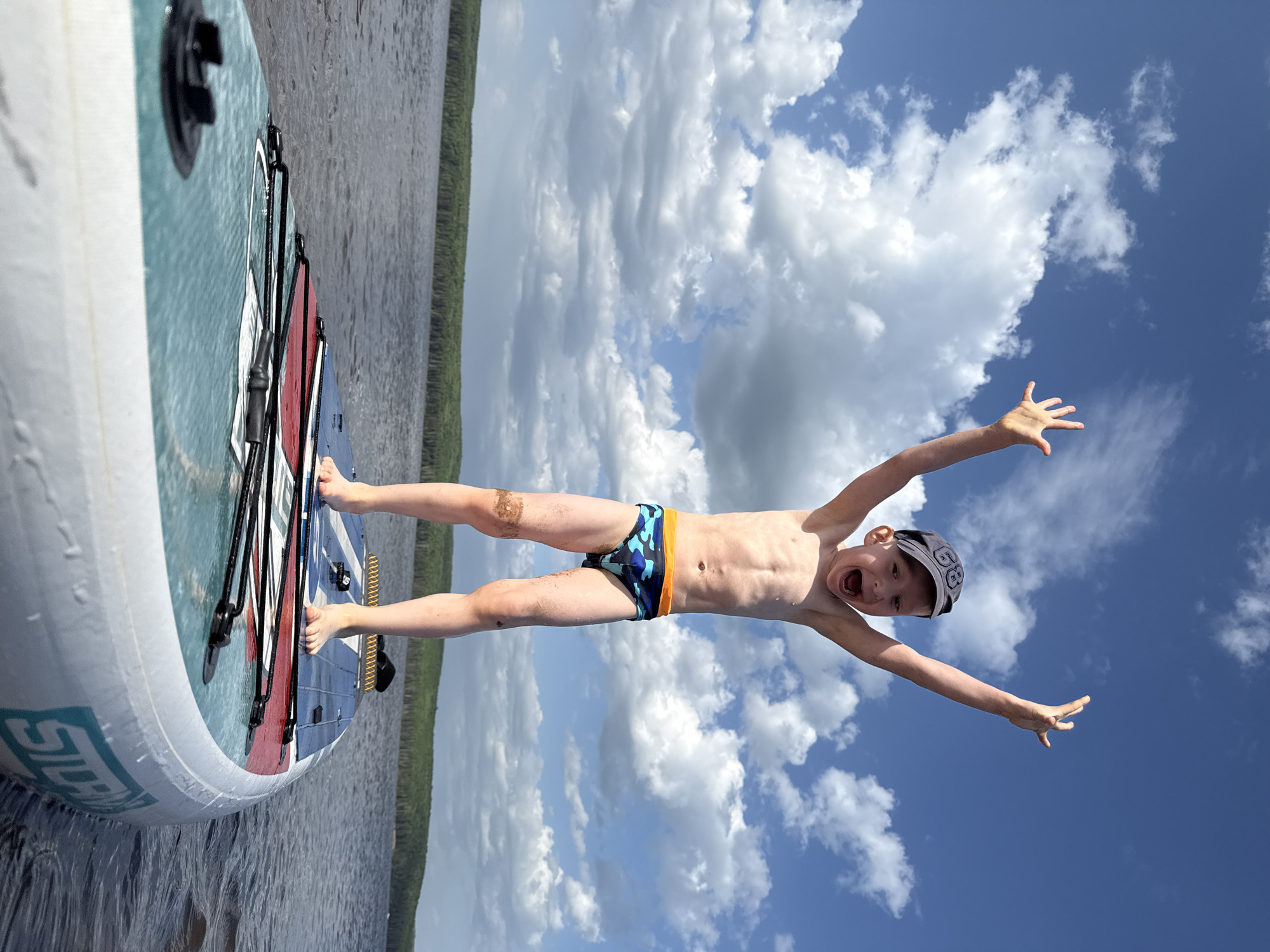 A boy wearing swim trunks and a baseball cap is lying on a paddleboard in Chaikovsky, Russia. He is facing upwards towards a partially cloudy sky. His arms are outstretched and he is yelling. The paddleboard is partially submerged in the water of what appears to be a lake or river. Trees are visible in the distance along the shoreline. The lighting indicates it is daytime.