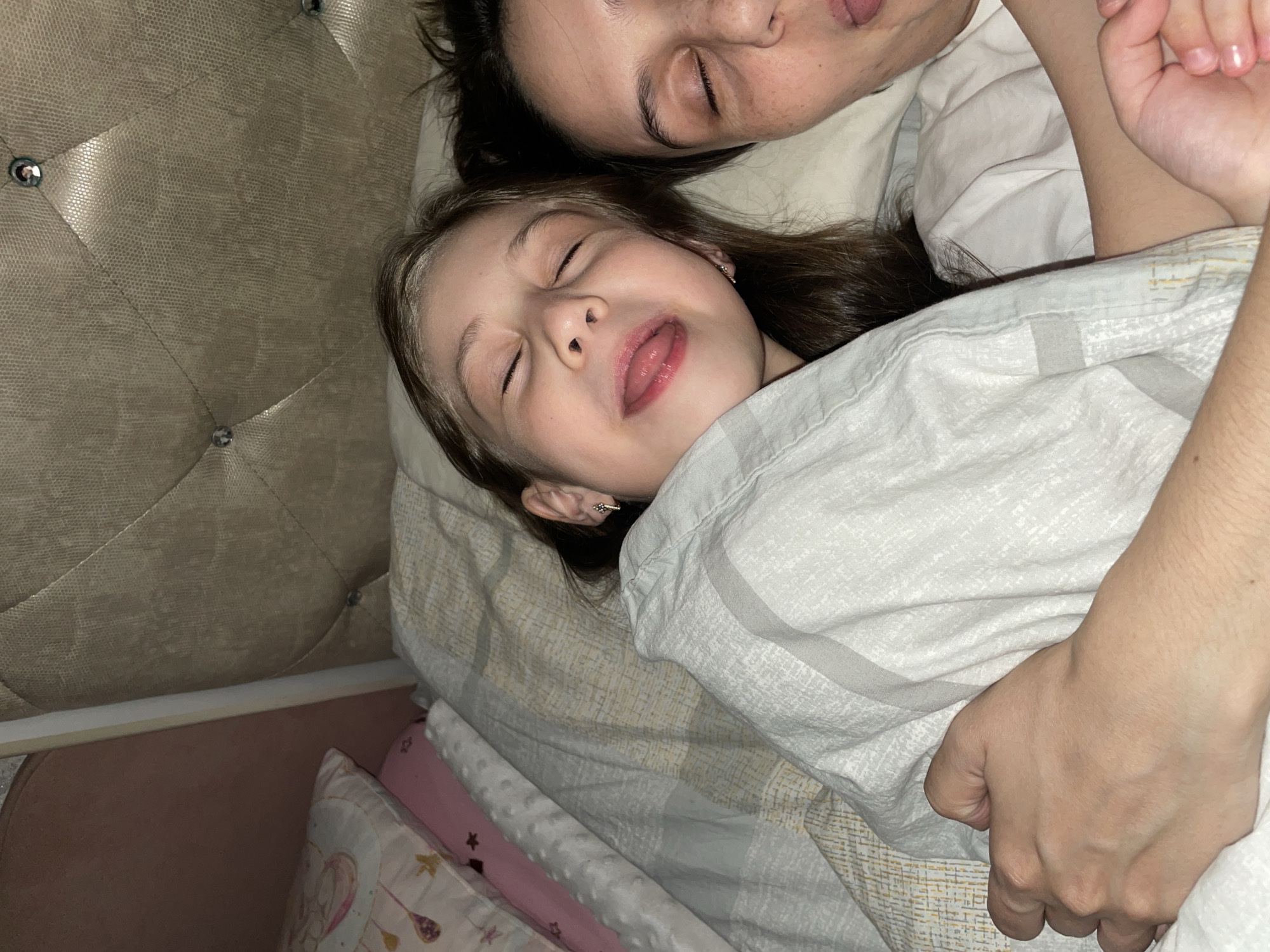 A young girl lies on a bed, eyes closed and tongue slightly protruding from her mouth. An adult person, also with closed eyes, is next to her. Both are covered by a patterned duvet, with an arm draped protectively over the girl. A headboard with diamond-shaped buttons is visible behind them. The scene appears to be indoors, and the specific location in Izhevsk, Russia, cannot be determined from the image.