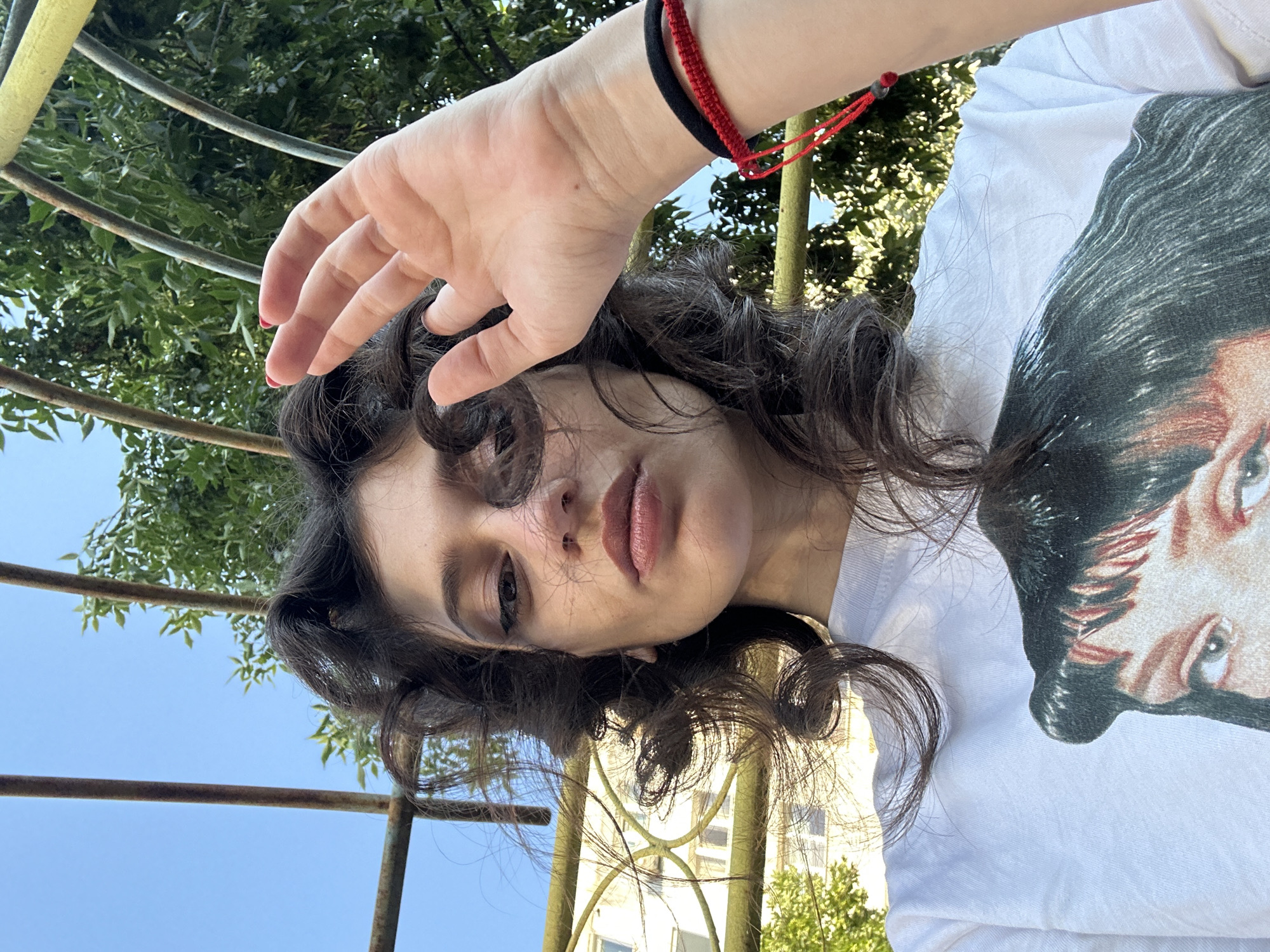 A young woman is posing for a selfie outdoors on a sunny day. She has dark, curly hair that frames her face and is looking towards the camera with a neutral expression. Her right hand is raised, obscuring part of her face and the background. She is wearing a white t-shirt with a graphic print of a woman's face on it. On her right wrist, she is wearing two bracelets, one black and one red. Behind her, there is a metal structure that appears to be part of a playground or park bench, with green foliage and a clear blue sky visible above. A building with windows is visible in the background, suggesting an urban setting. The scene is lit by natural sunlight, creating shadows and highlights on the woman's face and arm. The photograph appears to have been taken from a low angle.