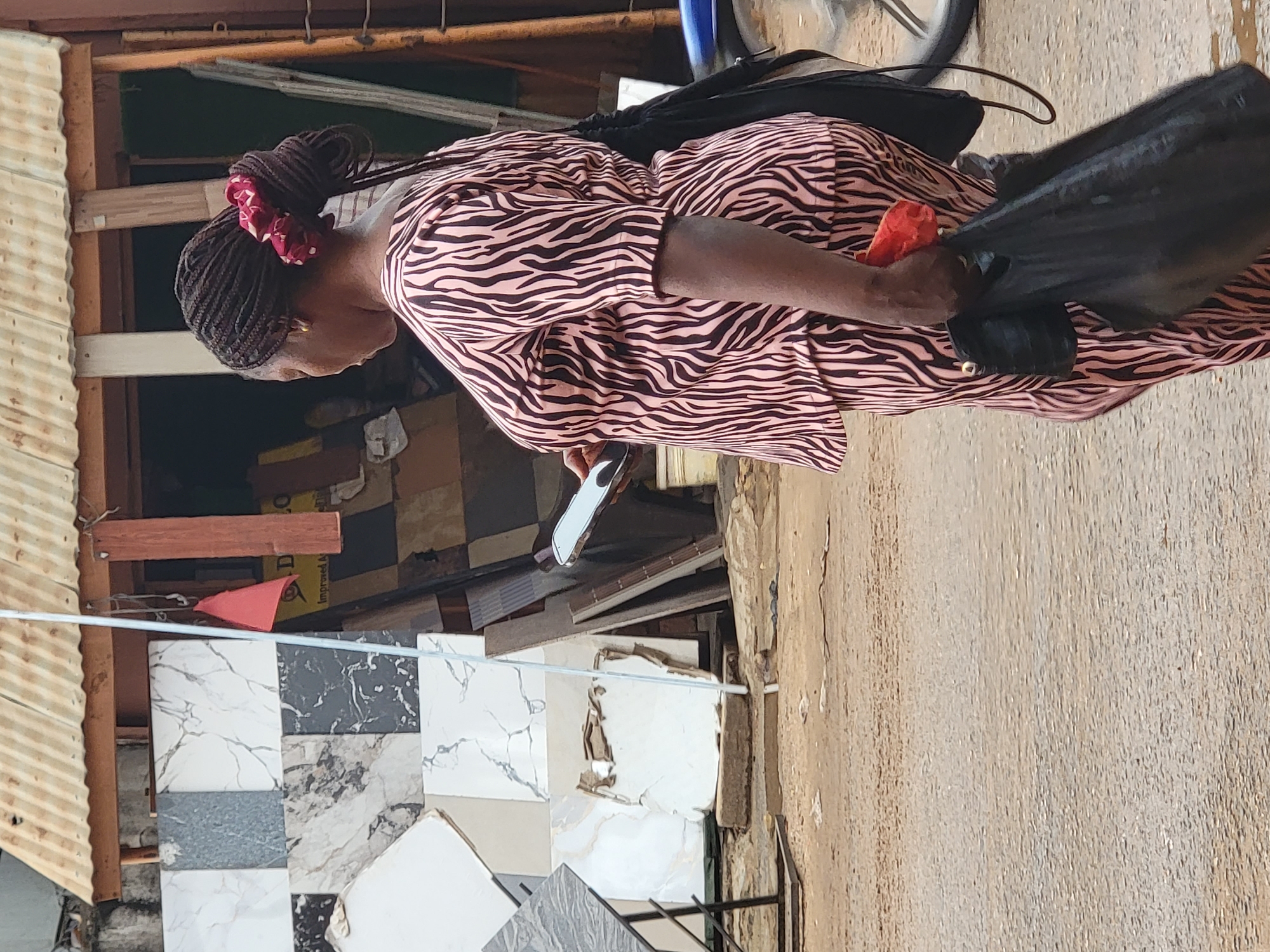 The image, taken in Maiburiji, Nigeria, captures a woman walking along an outdoor street or market area during what appears to be a bright, clear day. The photograph is oriented sideways, showing the woman in profile, moving from right to left across the frame if rotated correctly.
The main subject is a woman with dark skin and dark braided hair, styled into an elaborate bun or updo at the back of her head, adorned with a prominent red floral hair tie. She is wearing a distinctive pink and black zebra-print dress or tunic. A gold earring is visible in her left ear. She holds a silver smartphone in her left hand, which is down by her side. In her right hand, held closer to her body, she carries a black bag and a small, bright red object, possibly a piece of fruit or fabric.
The setting appears to be a bustling commercial or semi-urban environment. To the left, various patterned floor tiles, including marble-effect and plain grey designs, are displayed vertically, suggesting a tile vendor or building materials store. Corrugated metal sheeting and wooden planks are visible behind these tiles, forming part of a structure. A thin white cable or string stretches horizontally across the mid-ground. In the upper right corner, part of a bicycle wheel can be seen.
Visible text in the background is fragmented; on a cardboard box, parts of words like "D LO", "Improved A...", and "Internet d..." are discernible. The ground beneath the woman's feet is a light, textured surface, typical of an unpaved or lightly paved street. The overall impression is of daily life unfolding in a vibrant, sunlit outdoor space.