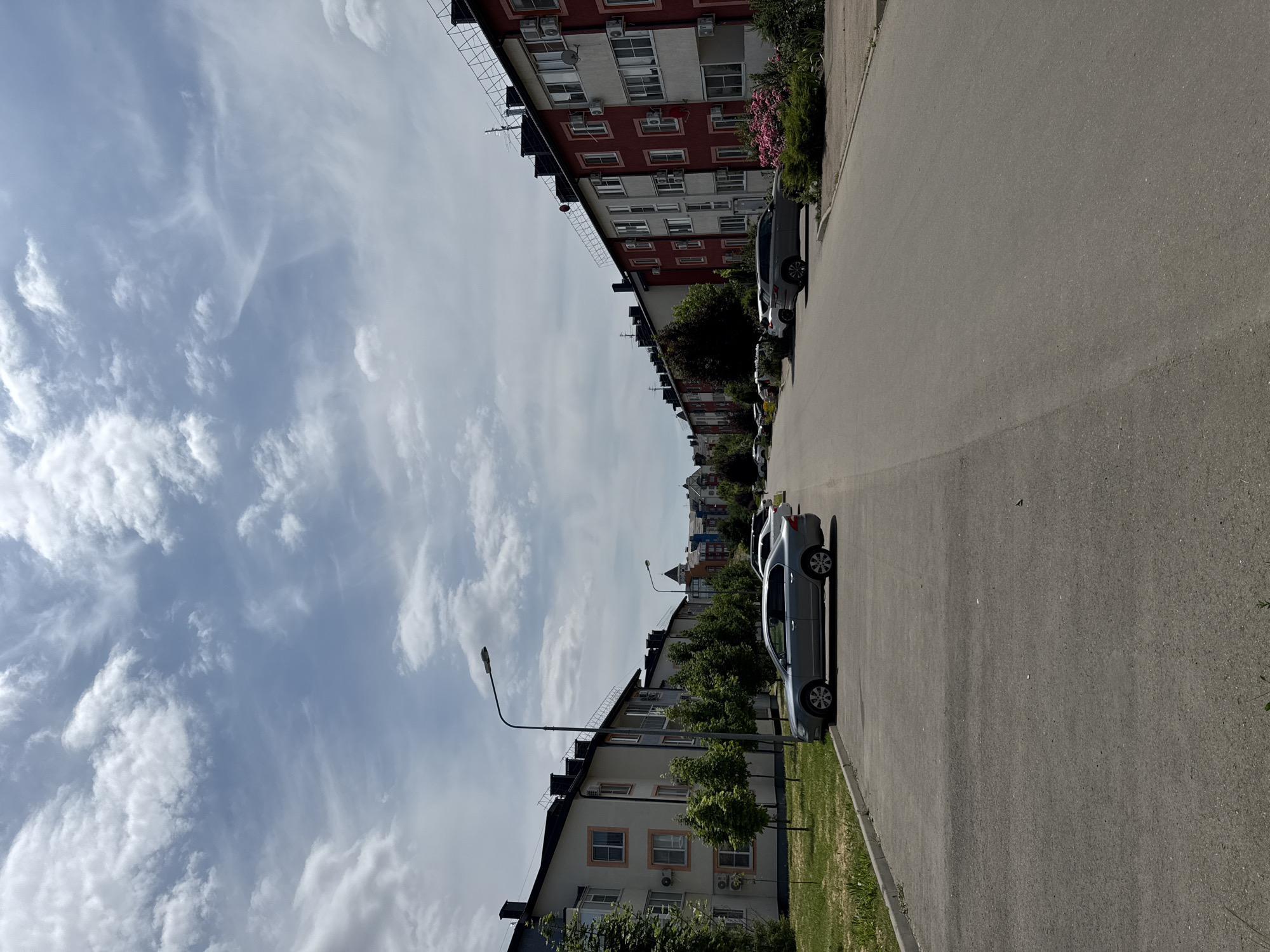 The image depicts a street scene in Krasnodar, Russia, likely a residential area characterized by multi-story buildings on either side of a paved road. The architecture of the buildings varies, with some featuring light-colored facades and dark roofs, while others are painted in bolder colors like red and white. Several cars are parked along the street, including a silver sedan in the foreground and an SUV further down. Green trees and manicured lawns line the sidewalks, suggesting a well-maintained urban environment. The sky is partly cloudy with scattered white clouds against a blue background. The overall impression is a calm and ordinary day in a suburban or residential district of Krasnodar.