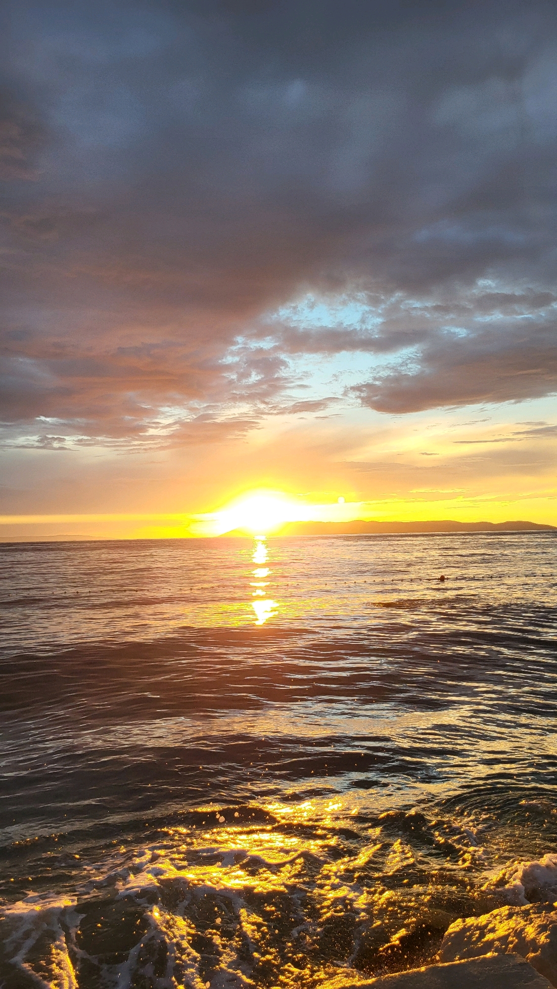 This image depicts a sunset over the Adriatic Sea in Općina Tučepi, Croatia. The sun is low on the horizon, casting a golden glow on the water. The sky is a mix of dark clouds and lighter hues of orange and yellow. Gentle waves are visible in the foreground, with some white foam. The horizon line is marked by distant landmasses. There are no people or man-made objects clearly visible in the scene, apart from a faint line of buoys or markers in the distance on the water. The overall impression is one of natural beauty and tranquility.