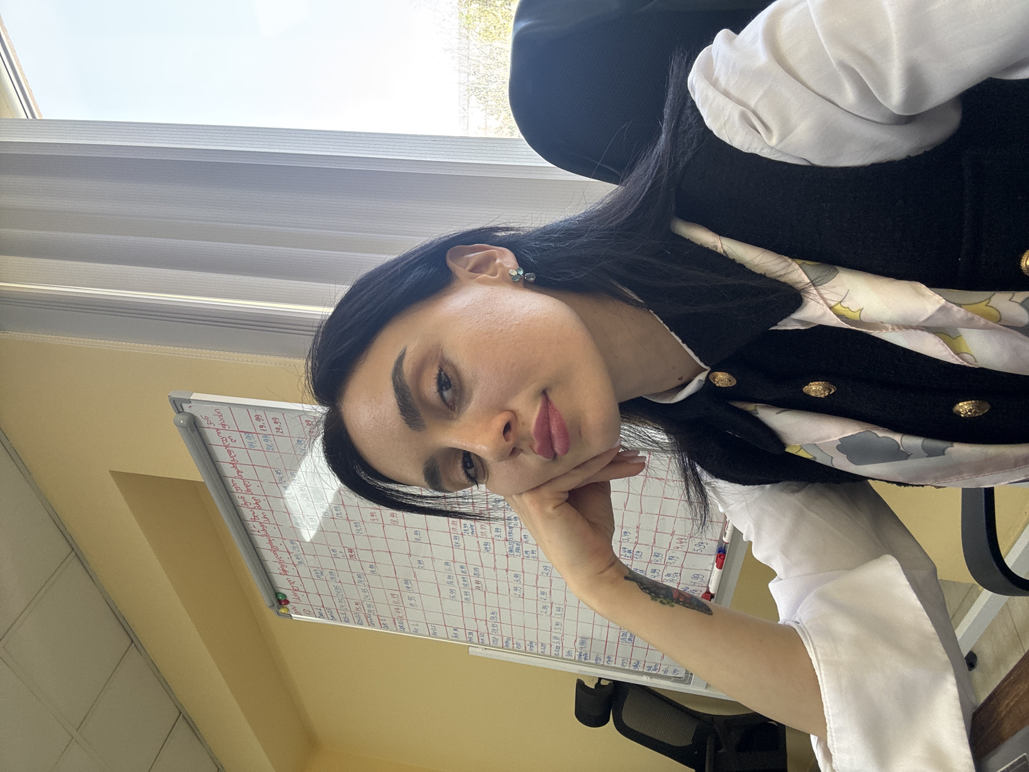 A young woman is captured in a selfie-style shot, leaning her head on her hand and looking directly at the camera with a slight smile. She has dark hair, dark eyebrows, and is wearing a black vest or jacket with gold buttons over a white blouse and a light-colored, patterned scarf. A small earring is visible on her left ear, and a tattoo, possibly depicting a butterfly or moth, is visible on her right forearm.
The setting appears to be an office or workspace, with a large whiteboard dominating the background behind her. The whiteboard is filled with handwritten text, numbers, and grid lines. To her left, an office chair is partially visible. Behind her, a window with horizontal blinds lets in bright daylight, suggesting it is daytime with clear weather. Through the top of the window, a blue sky is visible, and trees are faintly seen through the side. The walls are a light yellow color.
Visible text on the whiteboard includes numerous numbers like "0.99," "1.99," "2.99," "3.99," "4.99," "6.99," "11.99," "14.99," "16.99," "18.99," "19.99," "24.95," "24.99," "29.99," and "37.99," suggesting price lists or inventory. Words like "Air," "AirOS," "Master," "KOS," and "υποστυφό" are also present. The writing appears to be in a language other than English, likely Georgian given the provided context.
