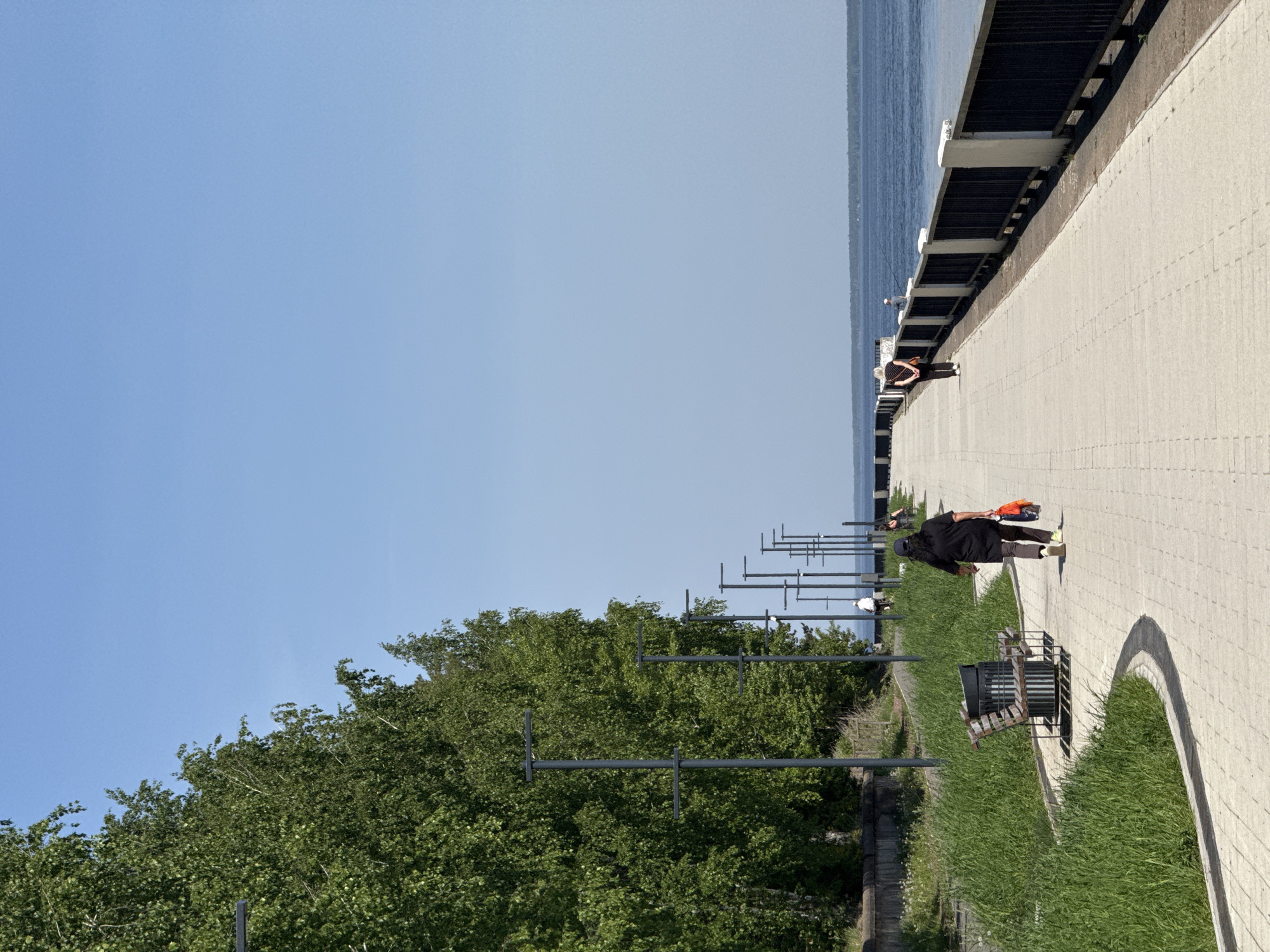 The image depicts a waterfront scene on a sunny day. A paved walkway stretches along the water's edge, bordered by a black metal railing on the water side. The walkway is textured with small, evenly spaced bricks. A person is walking along the walkway. Adjacent to the walkway, there is a strip of grass with a metal bench, extending to a line of tall green trees. Further back, utility poles stand along the waterfront. The sky is clear and blue. A second person is seen fishing along the waterway. The location is in the city of Unknown, Unknown.
