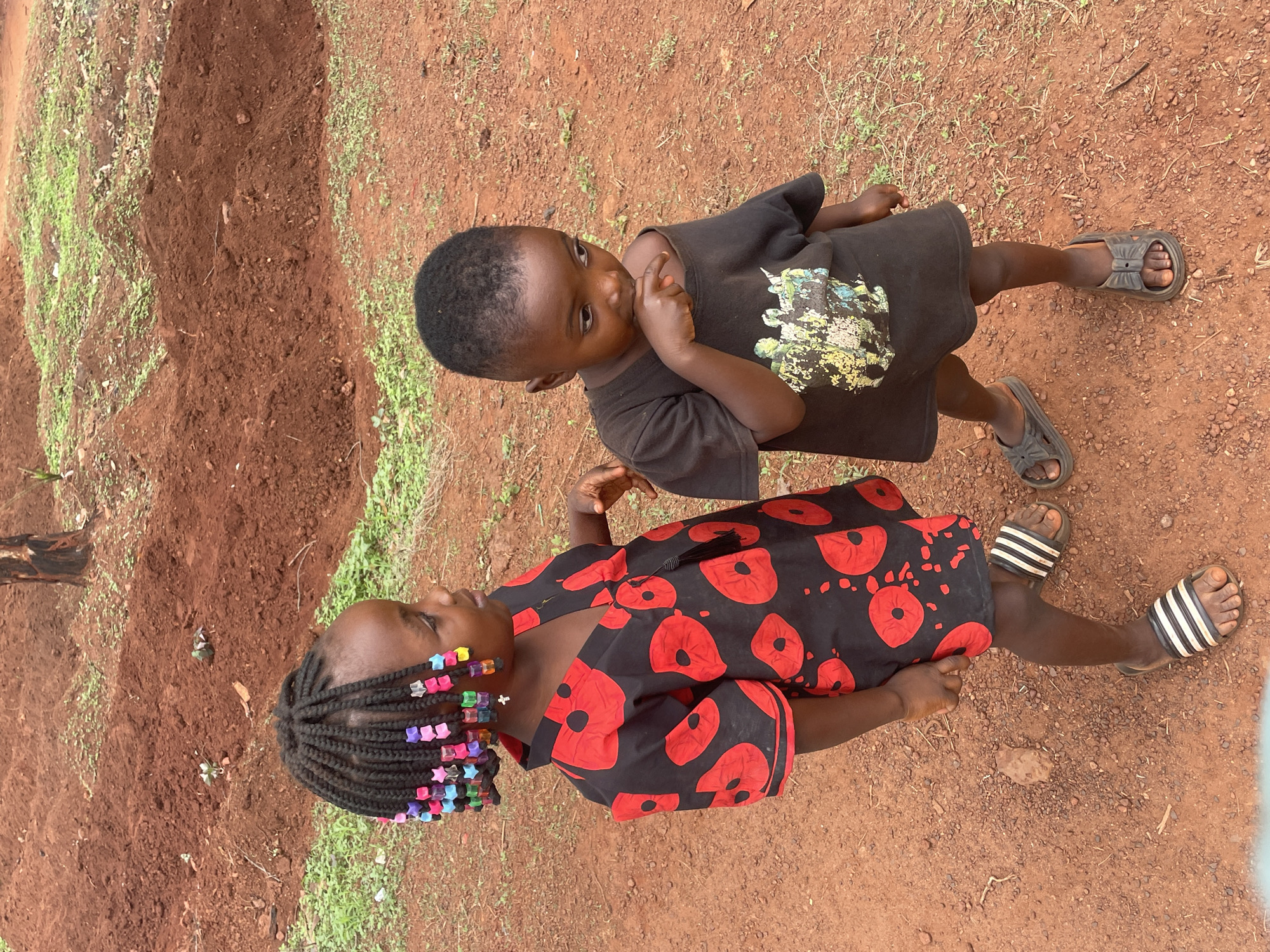 Two children stand on red earth ground in Ogbe, Nigeria. One child, facing the viewer, wears a black dress with large red poppy-like flowers, braids adorned with multicolored beads, and black and white striped sandals. The other child, partially obscured, wears a black t-shirt with a graphic design and gray sandals. The red earth ground is dry and contains small pebbles. Green vegetation is visible at the edges of the scene.
