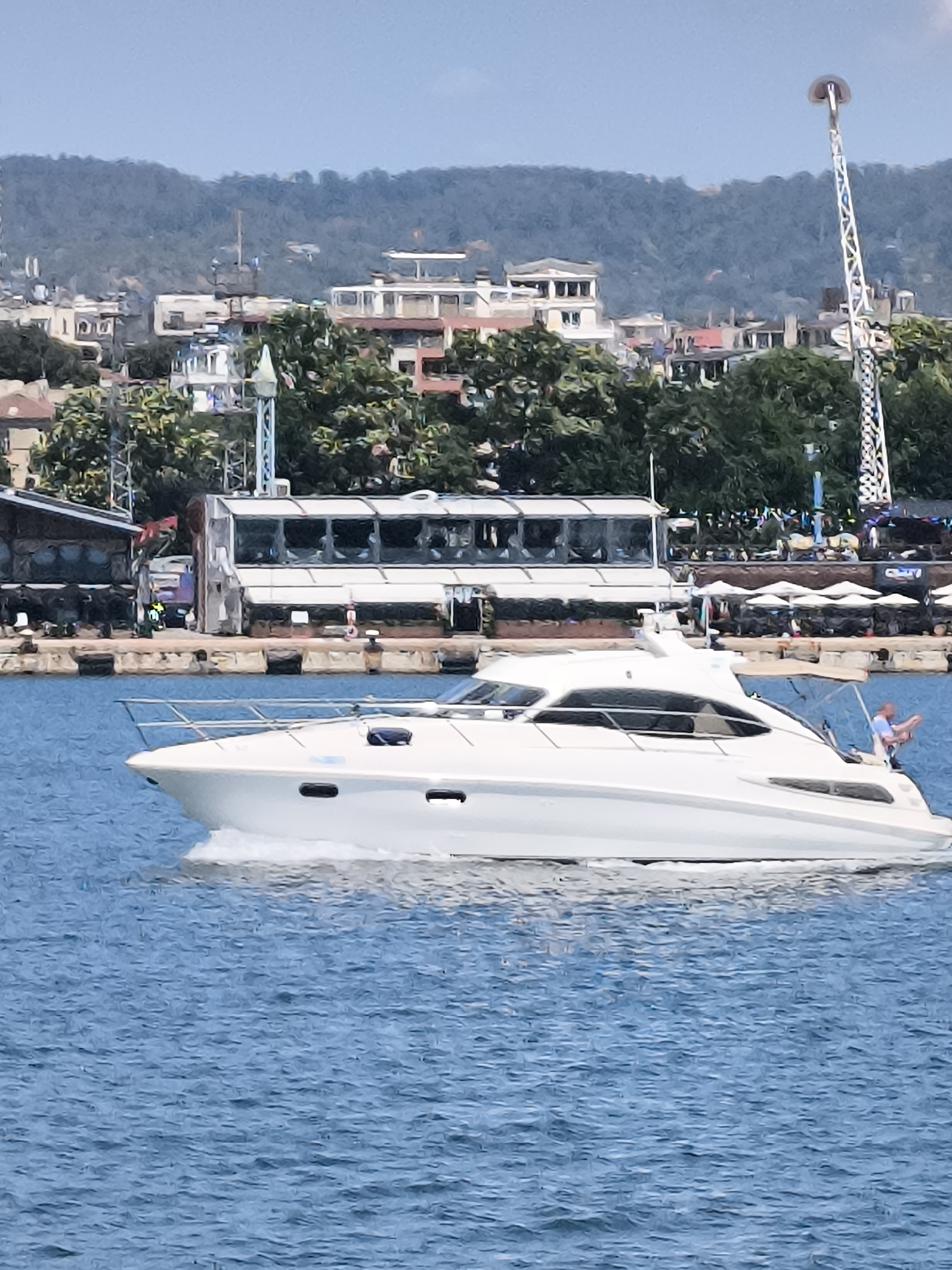 A white motor yacht is cruising on the blue sea in front of a coastal city. The yacht has a sleek design with a white hull and cabin. A person is visible on the stern of the yacht, sitting with their legs dangling over the side. In the background, the city of Varna, Bulgaria, is visible. The cityscape features a mix of buildings, trees, and a tall, slender amusement park ride. The water is choppy, with white foam trailing behind the yacht. The sky is a clear, pale blue.