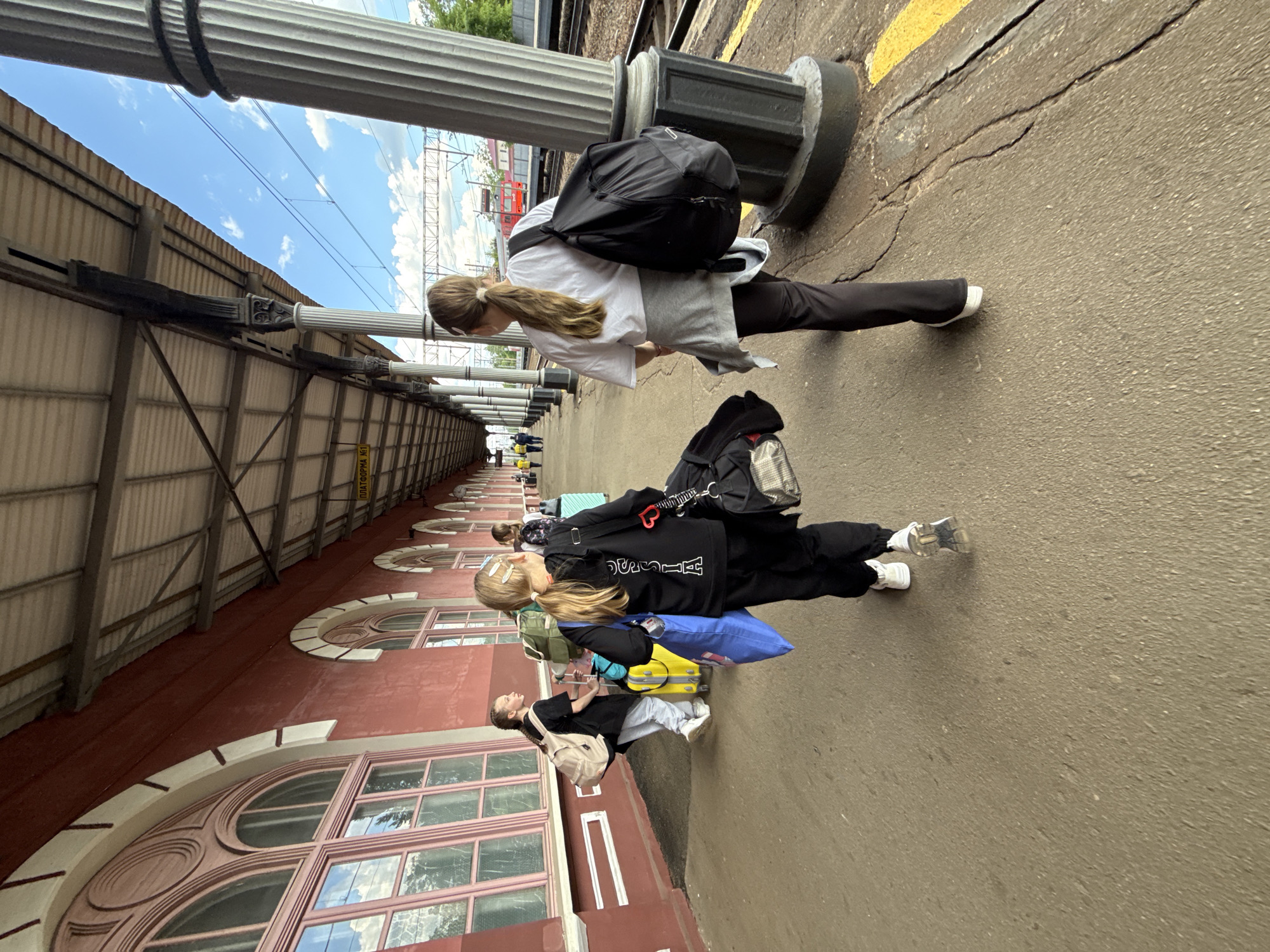 The image, taken in Malaya Vishera, Russia, depicts a group of people waiting on a train platform next to a historic-looking building, under a bright daytime sky. The photograph is rotated 90 degrees clockwise.
In the foreground, a young woman with long, light brown hair tied in a ponytail wears a white t-shirt, dark pants, and a black backpack, standing beside a large, ornate white column with a dark base. Behind her are two younger females, also with long hair. One is dressed in a black tracksuit with a black backpack, carrying a blue bag and pulling a yellow suitcase. The other wears a grey t-shirt, light-colored pants, and a beige backpack, and also has a yellow suitcase. Additional indistinct figures are visible further down the platform.
The train platform features cracked asphalt and a yellow safety line. To the left, a long, red-hued building with classical architectural elements, such as multiple large arched windows with decorative light-colored frames and details, stretches into the distance. Above the building's roof, which appears to be corrugated metal, a bright blue sky with scattered white clouds is visible. Ornate white columns support an overhead structure or canopy above the platform. In the background, railway tracks and a red train or wagon are partially visible. A yellow sign is partially visible in the distance, reading "ПЛАТФОРМА НО1" (PLATFORM NO1). The overall scene suggests travelers waiting for a train in pleasant weather conditions.