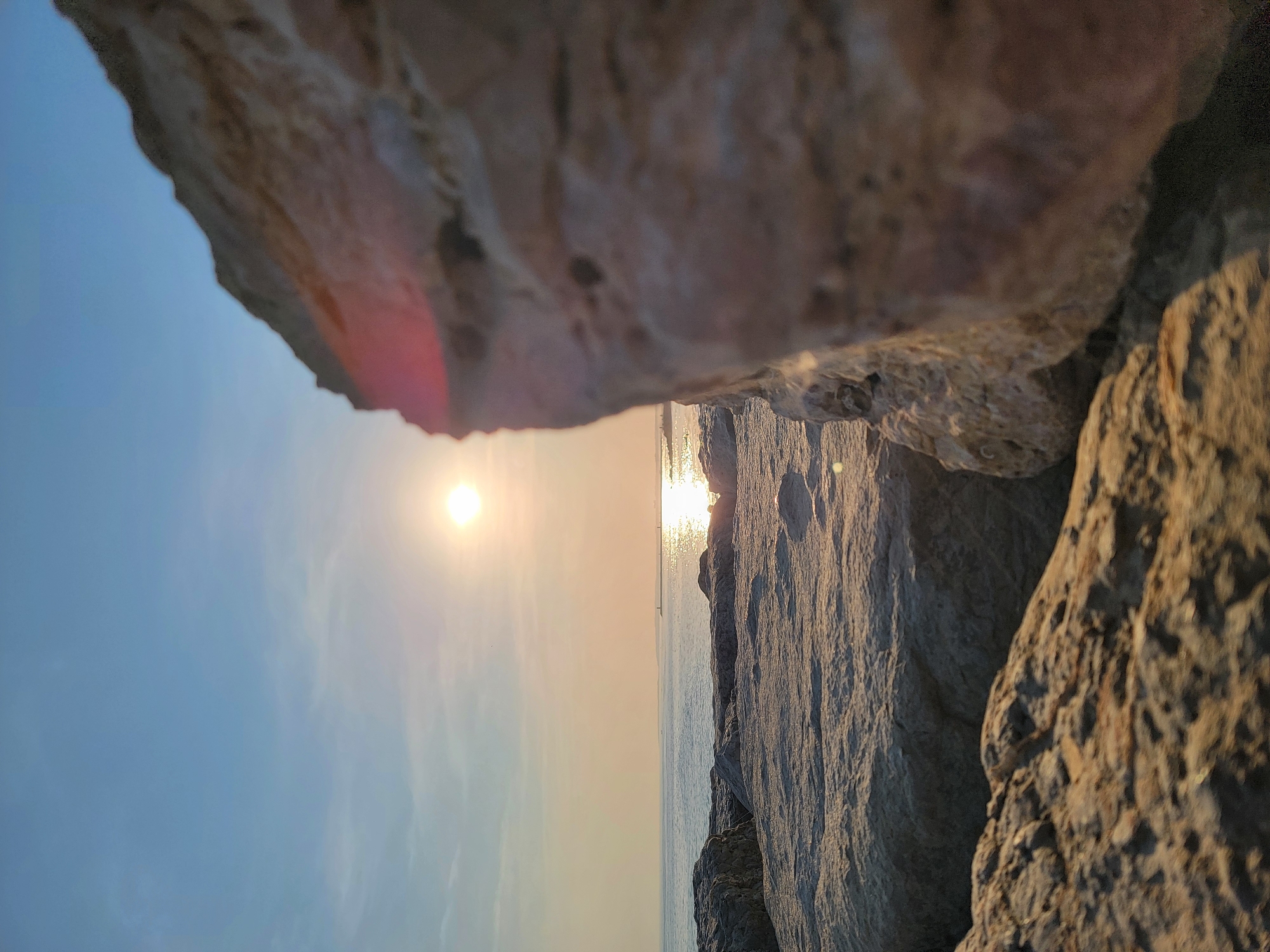 The image captures a sunset scene at the Quarteira, Portugal coastline. In the foreground, large, textured rocks, likely part of a breakwater or natural rock formation, dominate the lower portion of the frame. The rocks are a light beige and brown color, with visible pores and rough surfaces, catching the warm light of the setting sun.
The middle ground features the calm, reflective surface of the ocean, with the sun's glare creating a shimmering path across the water. The horizon line is visible, with a faint outline of land or structures in the distance. The sky transitions from a pale yellow near the horizon to a clear, soft blue at the top of the image, with subtle wisps of clouds.
A significant portion of the right side of the image is taken up by a large, out-of-focus rock that creates a strong foreground element and partially obscures the view of the sea and sky. The overall lighting suggests late afternoon or early evening, with the sun positioned low in the sky. No people or specific urban elements are discernible in the immediate scene, focusing instead on the natural landscape and the interplay of light and shadow on the rocks and water.
