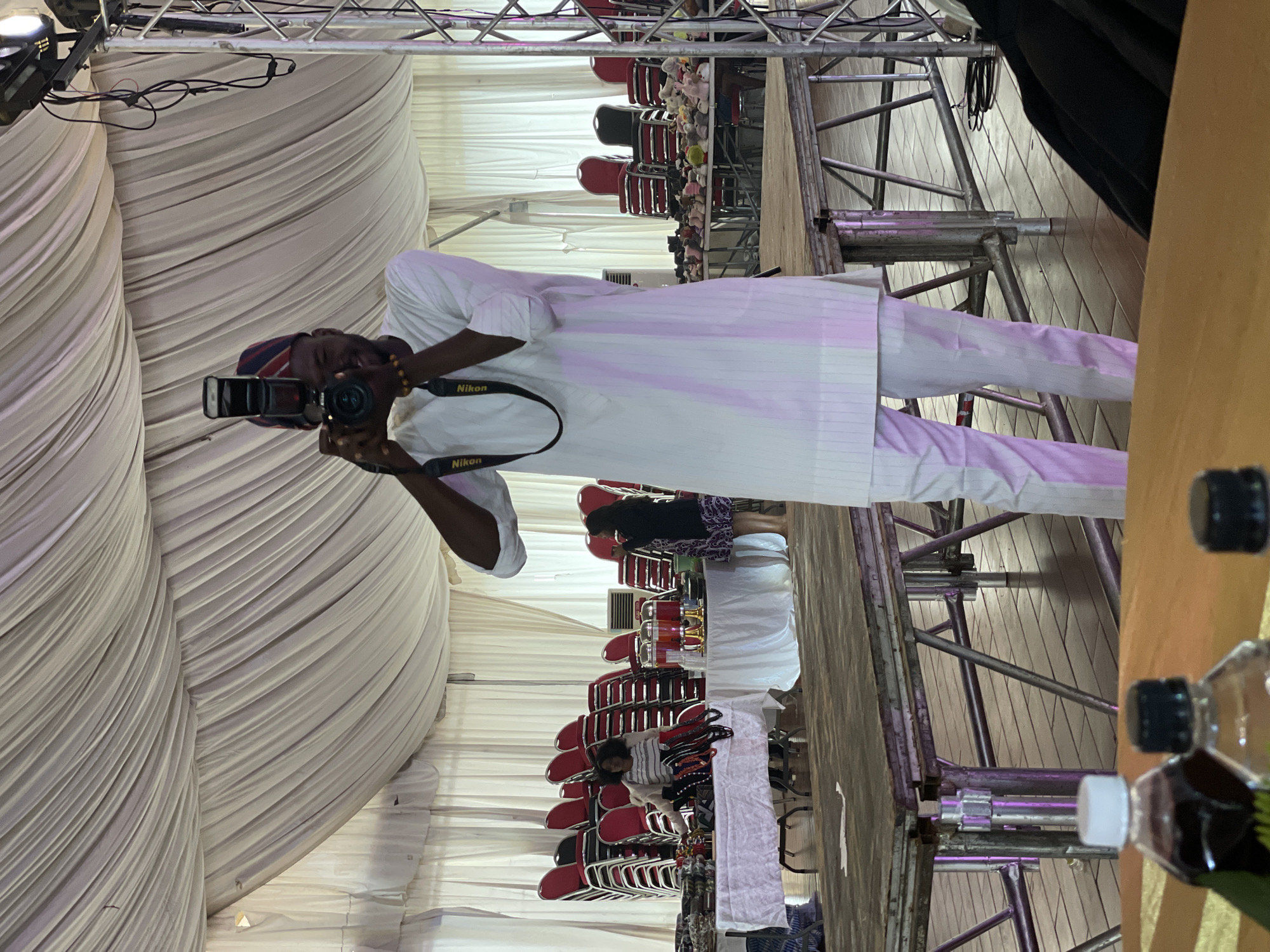 This is a vertically oriented, reflected image of a person holding a camera and taking a picture of their reflection. The subject is a person dressed in a white traditional outfit with purple pinstripes. They are wearing a red and blue patterned headband. The camera appears to be a Nikon DSLR, with a black lens and strap.
The scene is set inside a large tent with draped white fabric walls. In the background, there are rows of stacked red chairs. On a table behind the chairs, there are several bottles, some with red liquid. There are also some stuffed animals on the chairs.
The reflection shows a stage or platform structure made of metal scaffolding. The floor appears to be tiled. The lighting suggests it is daytime, likely indoors. The overall atmosphere appears to be related to an event or gathering. The person in the reflection seems to be engaged in documenting the event.
There is visible text on the camera strap: "Nikon". There is also text on the camera body, but it is illegible. The context provided indicates the location is Barakin Lerawi, Nigeria.
