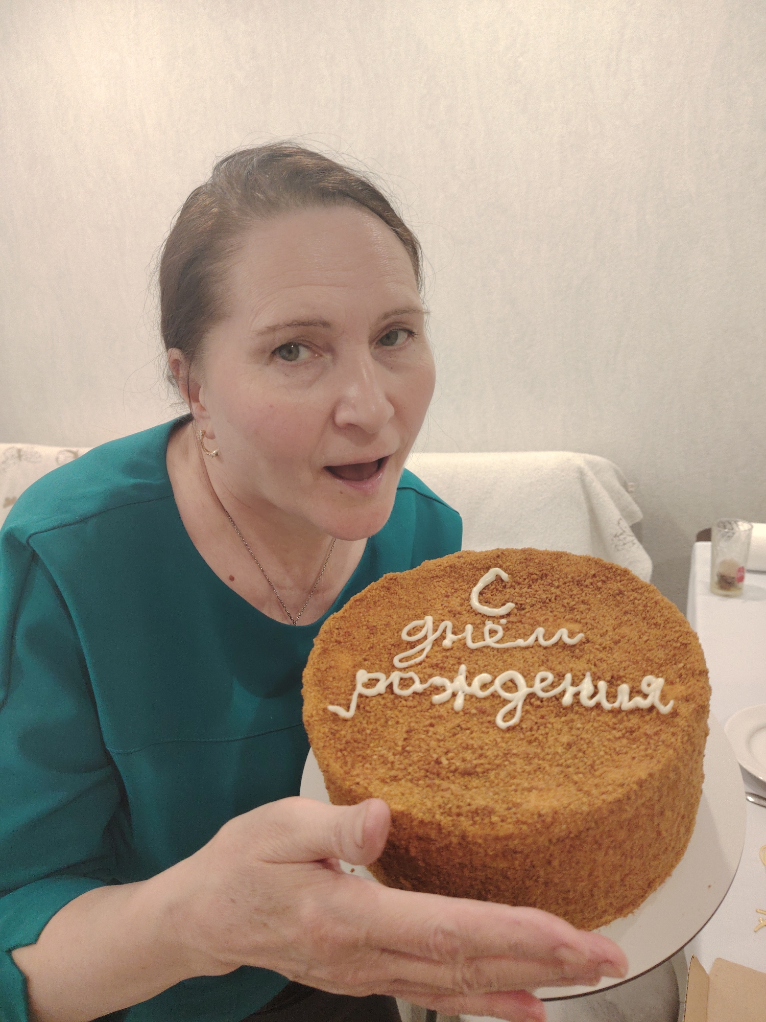 A woman holds a cake with the Russian phrase "С днем рождения" (Happy Birthday) written on top. She is wearing a green top, earrings, and a necklace. The cake is round and appears to be a honey cake ("Medovik"). The background includes a light-colored wall and a partially visible couch. The location is Sarapul, Russia.