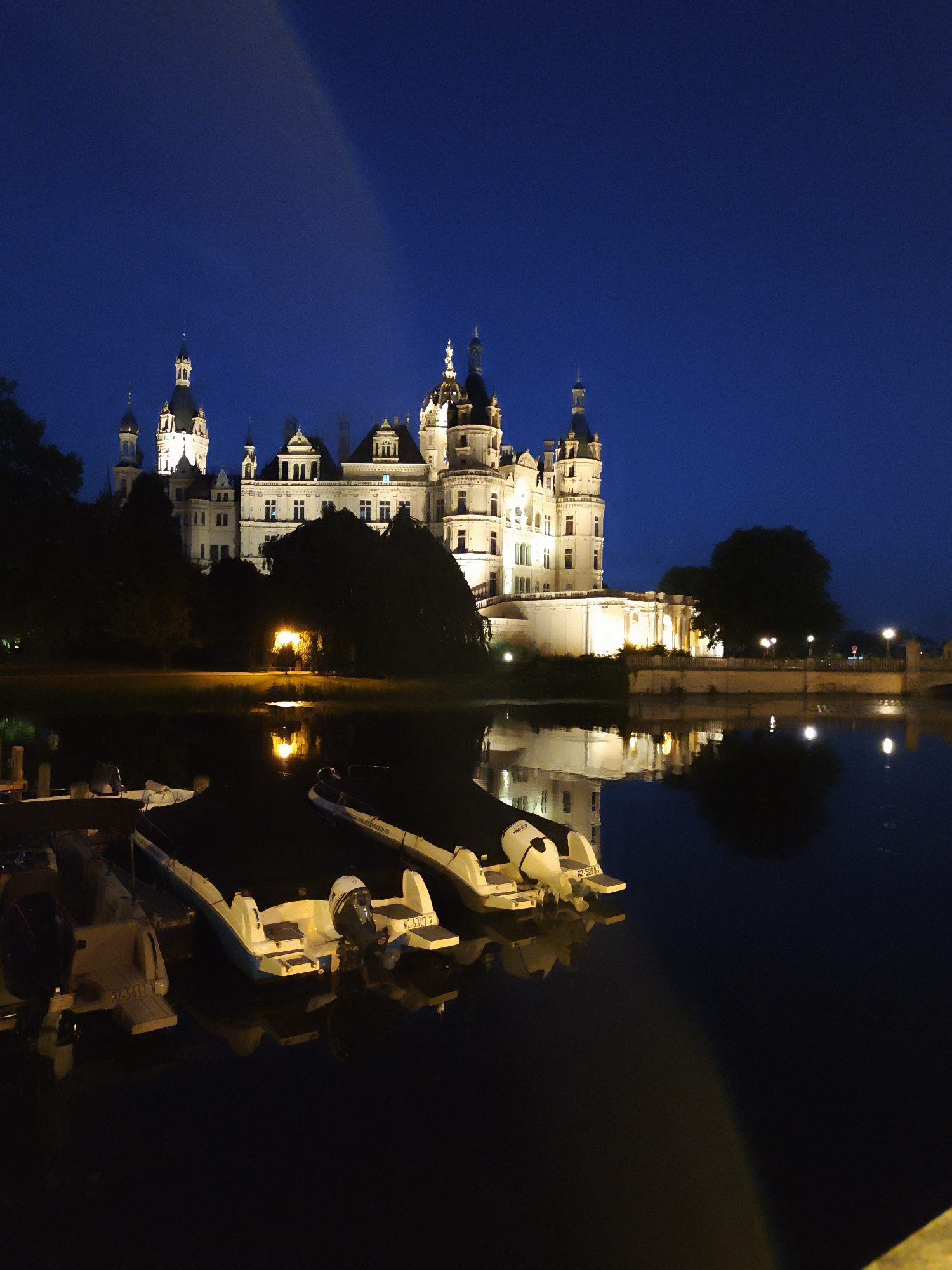 The image depicts a nighttime view of Schwerin Castle, a prominent landmark located on an island in the city of Schwerin, Germany. The castle is illuminated, highlighting its intricate architecture, which features multiple towers and spires. In the foreground, several small boats are docked on a body of water, likely the Schweriner See. The water reflects the lights from the castle and the boats, creating a serene and picturesque scene. The sky is dark blue, indicating it is nighttime. There are no people visible in the image.