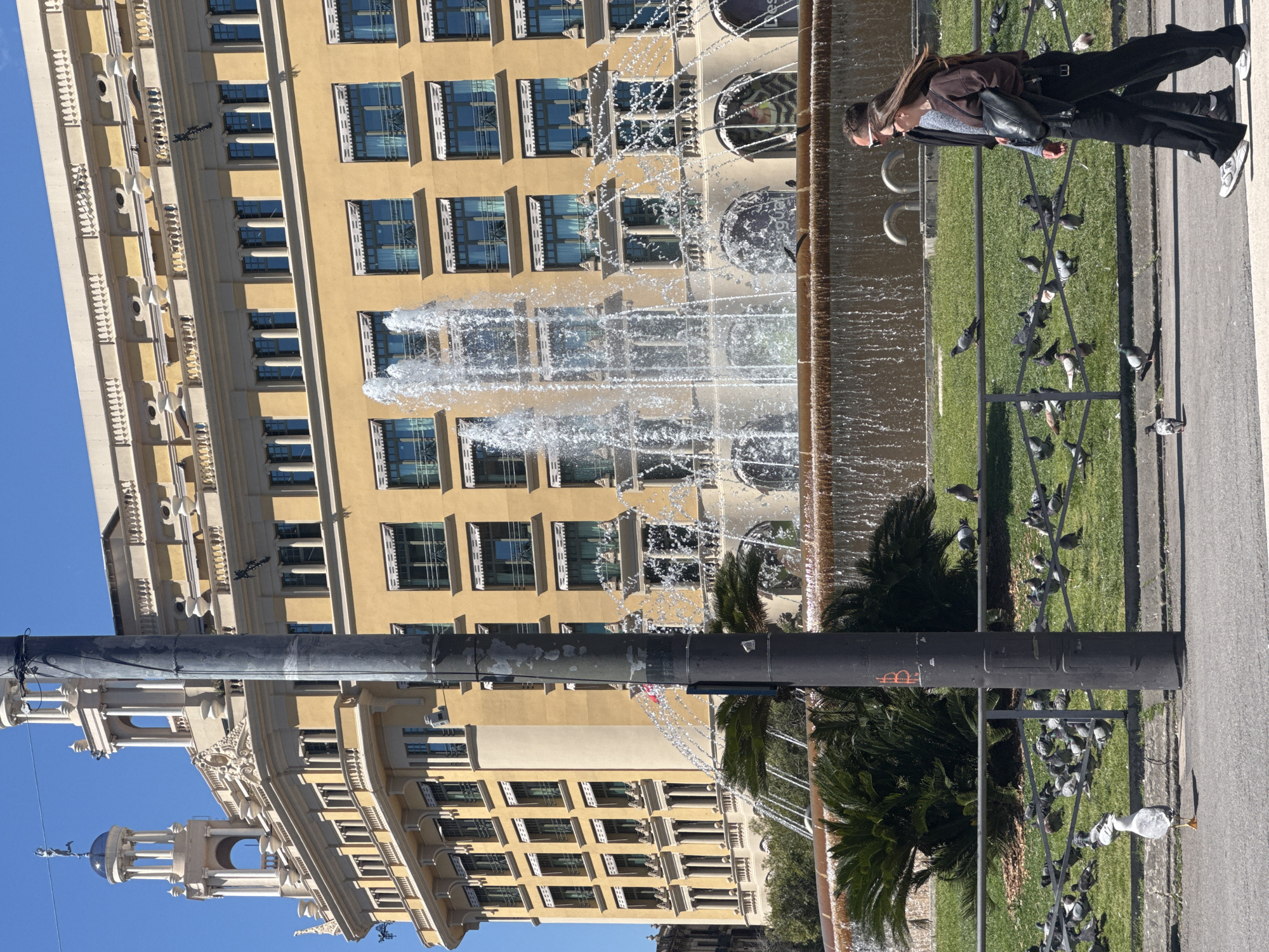 The image shows a street scene in Barcelona, Spain. A large, ornate building with multiple floors and windows stands in the background. In the foreground, a fountain with water spraying upwards is visible. A woman is standing near the fountain, seemingly observing the scene. Pigeons are gathered on the ground near a grassy area enclosed by a metal fence. A dark pole runs vertically through the frame, partially obscuring the view. The sky is blue.
