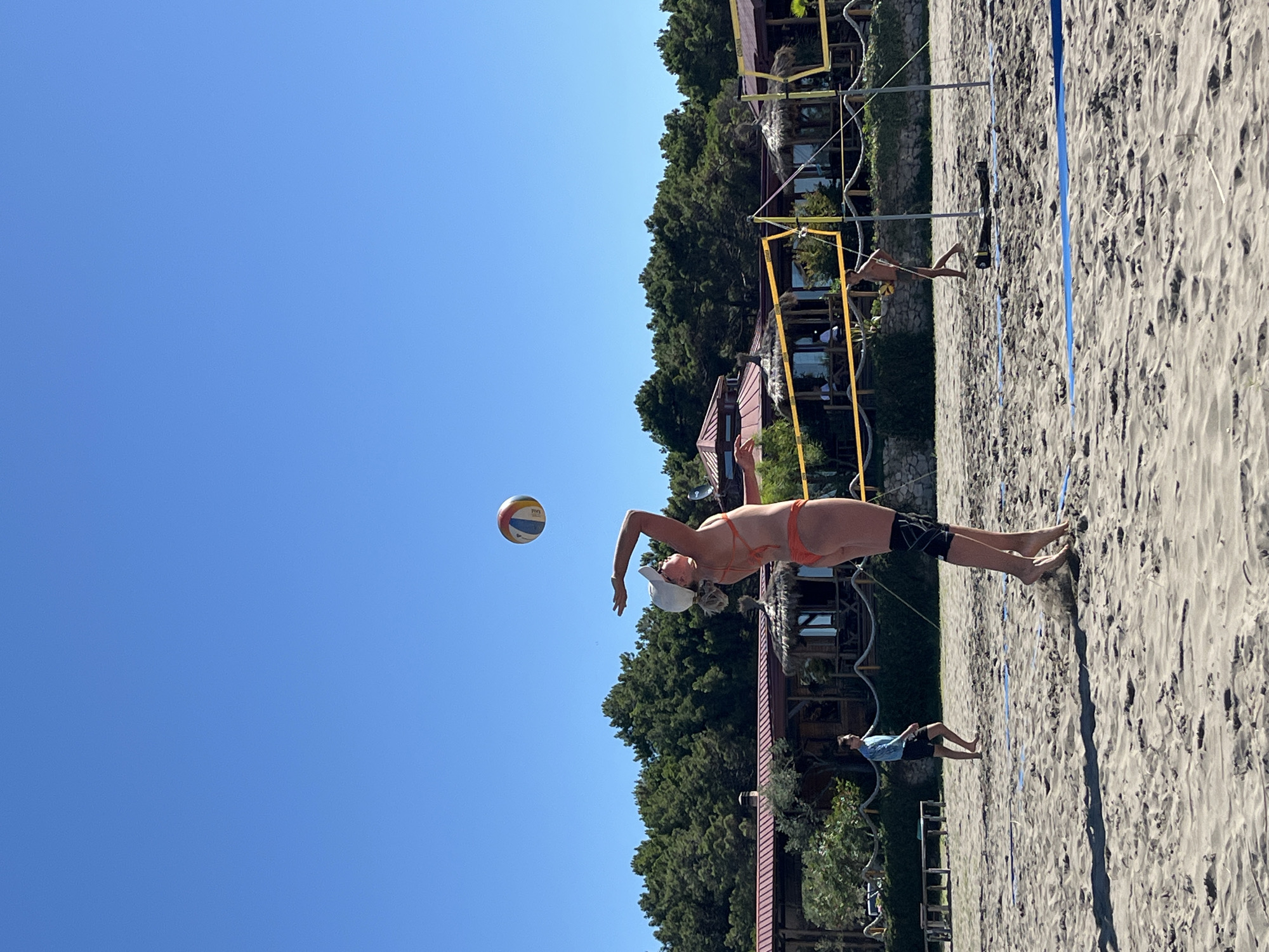 A group of people are playing beach volleyball on a sandy court. A woman in an orange bikini and white cap is in the foreground, leaping to hit the ball. She wears black knee pads. Behind her, a man in a blue shirt and black shorts walks across the court. Further back, another man in dark shorts is visible near the net, holding a volleyball. The beach volleyball court is marked with yellow and blue lines. In the background, there is a building with a reddish-brown roof and thatched umbrellas, surrounded by lush green trees. The sky is a clear, bright blue. The scene appears to be set in a beach resort in the city of Pistula, Montenegro.
