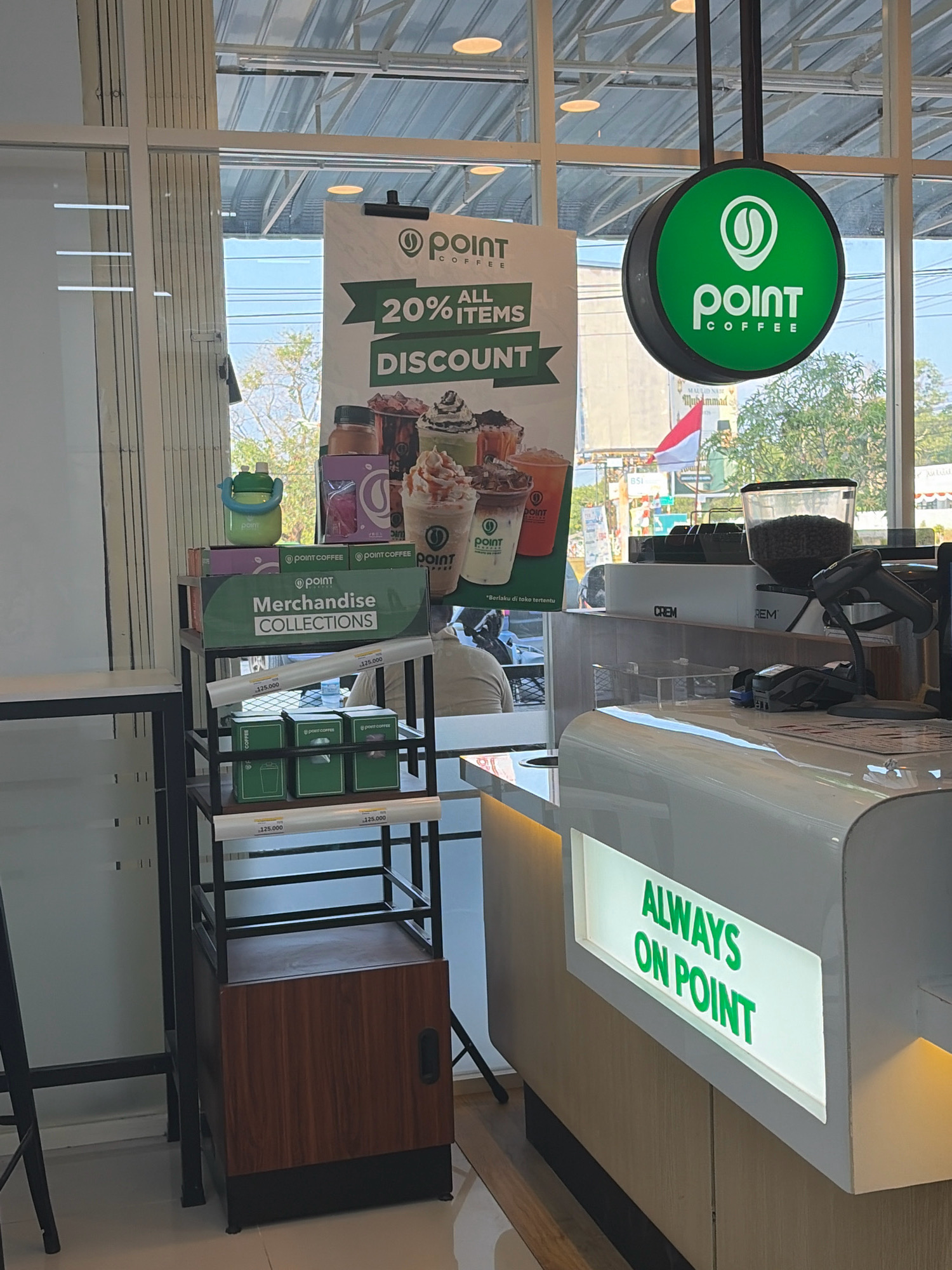 This image depicts the interior of a Point Coffee shop, a prominent chain in Indonesia, likely located in Tanjung Pinang. The scene is set within a modern retail environment characterized by clean lines and bright lighting. In the foreground, a white counter with a subtle illumination features a prominent LED sign displaying "ALWAYS ON POINT" in green lettering, reinforcing the brand's slogan. Behind the counter, a collection of coffee-making equipment is visible, including a grinder filled with coffee beans and a point-of-sale system with a barcode scanner.
To the left, a tiered display shelf showcases Point Coffee merchandise, including boxes of what appear to be coffee products and a branded water bottle. A large promotional banner hangs prominently, advertising "20% ALL ITEMS DISCOUNT," further emphasizing the brand's marketing efforts. The background reveals large windows offering a glimpse of the outside environment, which suggests a bright, sunny day with trees and buildings visible, consistent with the tropical climate of Tanjung Pinang. The overall impression is of a well-maintained and inviting coffee shop designed to attract customers and promote its products.