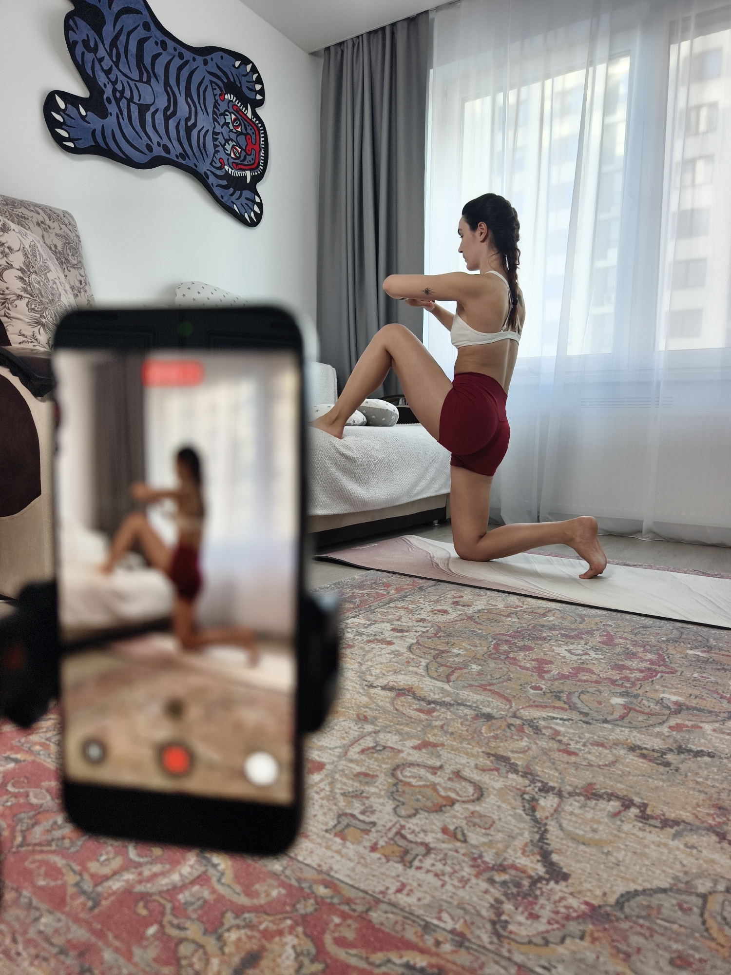 A woman in a white sports bra and red shorts is kneeling on a beige yoga mat indoors, twisting her upper body. She is being recorded by a smartphone set up in a holder in the foreground, which shows a blurred image of her on its screen. The scene is in a room with beige carpets, a bed in the background, a large blue tiger artwork on the wall, and a large window showing the cityscape outside. Based on the architectural style of the buildings visible through the window, the scene may be located in Tyumen, Russia.