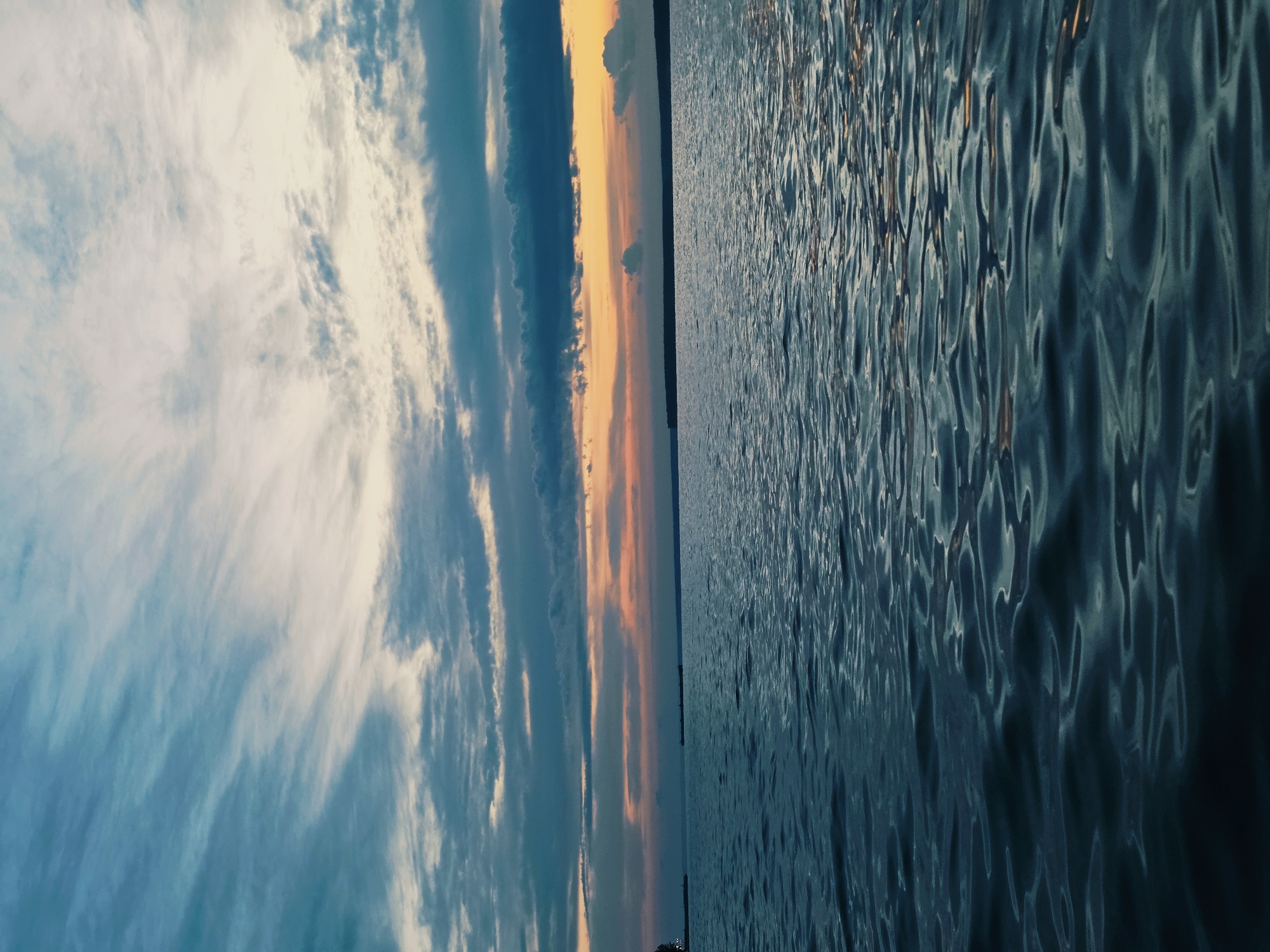 This image depicts a body of water, likely a river or lake, under a dramatic sky at dusk. The water's surface is rippled, reflecting the muted colors of the sky. The sky is filled with wispy clouds in shades of blue, gray, and orange, suggesting the sun has recently set or is about to rise. In the distance, a dark, low-lying horizon line separates the water from the sky, indicating land. There are no discernible people or specific landmarks in the foreground or midground. Given the information about Chaikovsky, Russia, this scene could be a view of the Kama River or a reservoir in the vicinity, a common natural setting in the region during twilight hours. The overall atmosphere is tranquil and serene.