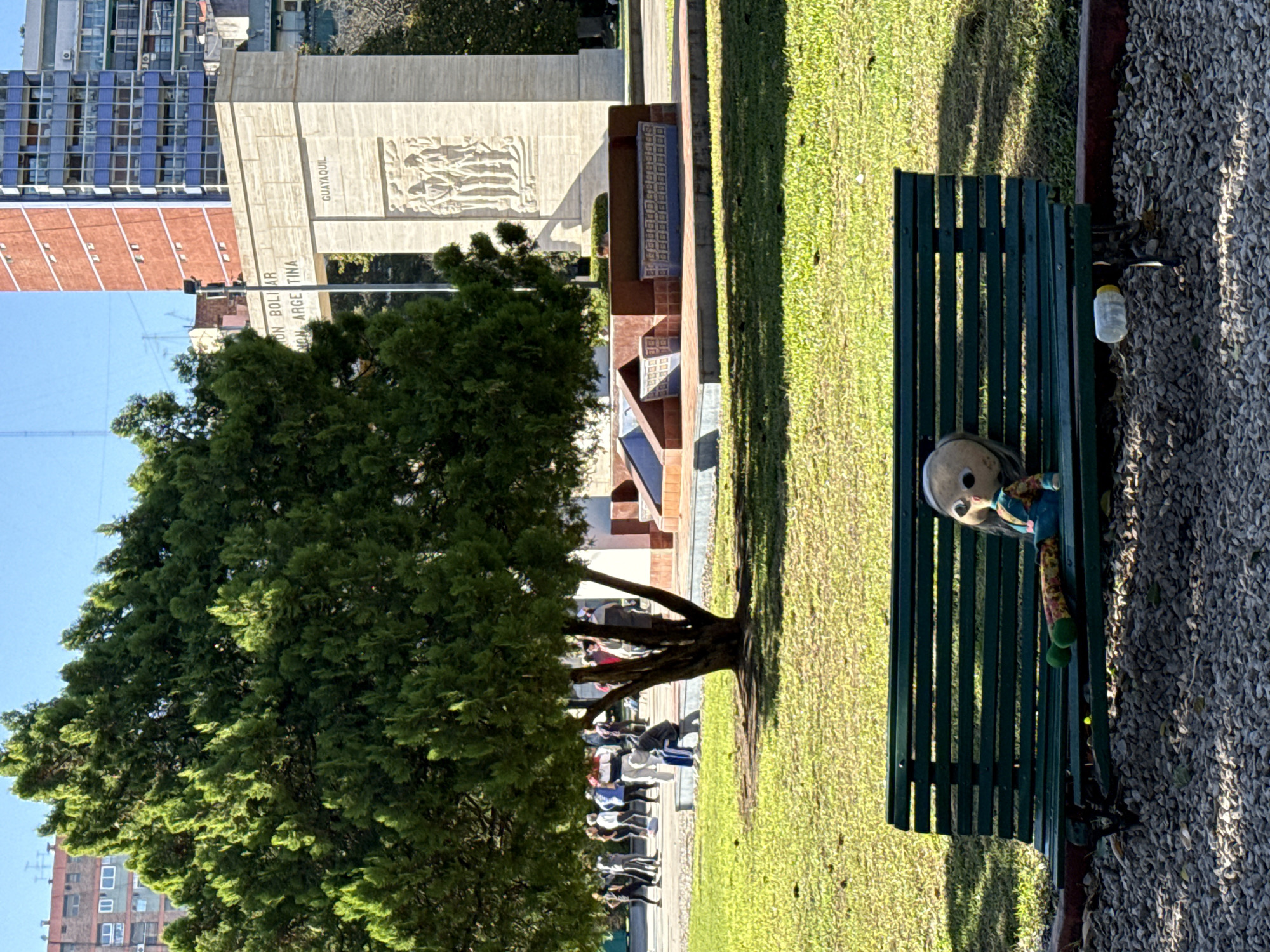 The image depicts a sunny day in a public park or plaza in Buenos Aires, Argentina. The scene is viewed from a slightly elevated position, looking across a green space towards a monument and city buildings.
In the foreground, a dark green slatted park bench sits on a patch of short green grass, bordered by a gravel pathway to its right. A light-skinned doll with bright clothing and socks, appearing to be a rag doll, is positioned on the bench. Next to the bench, on the gravel, a clear plastic bottle with a yellow cap is visible.
To the left, a large, dense evergreen tree with abundant foliage provides shade, and several people are seated on benches beneath its branches further in the background, appearing to be resting or socializing.
In the mid-ground and background, a large, light-colored concrete monument stands prominently. Carved reliefs depicting figures are visible on its side. Inscriptions on the monument read "SIMON BOLIVAR," "NACION ARGENTINA," and "GUAYAQUIL," with the year "1813" also present. Behind the monument, various city buildings rise, including one with blue-paned windows and another with reddish-brown brickwork.
The clear sky and strong shadows indicate it is daytime, likely morning or afternoon, with bright sunshine. The overall atmosphere appears peaceful and calm, characteristic of a public space being enjoyed by visitors.