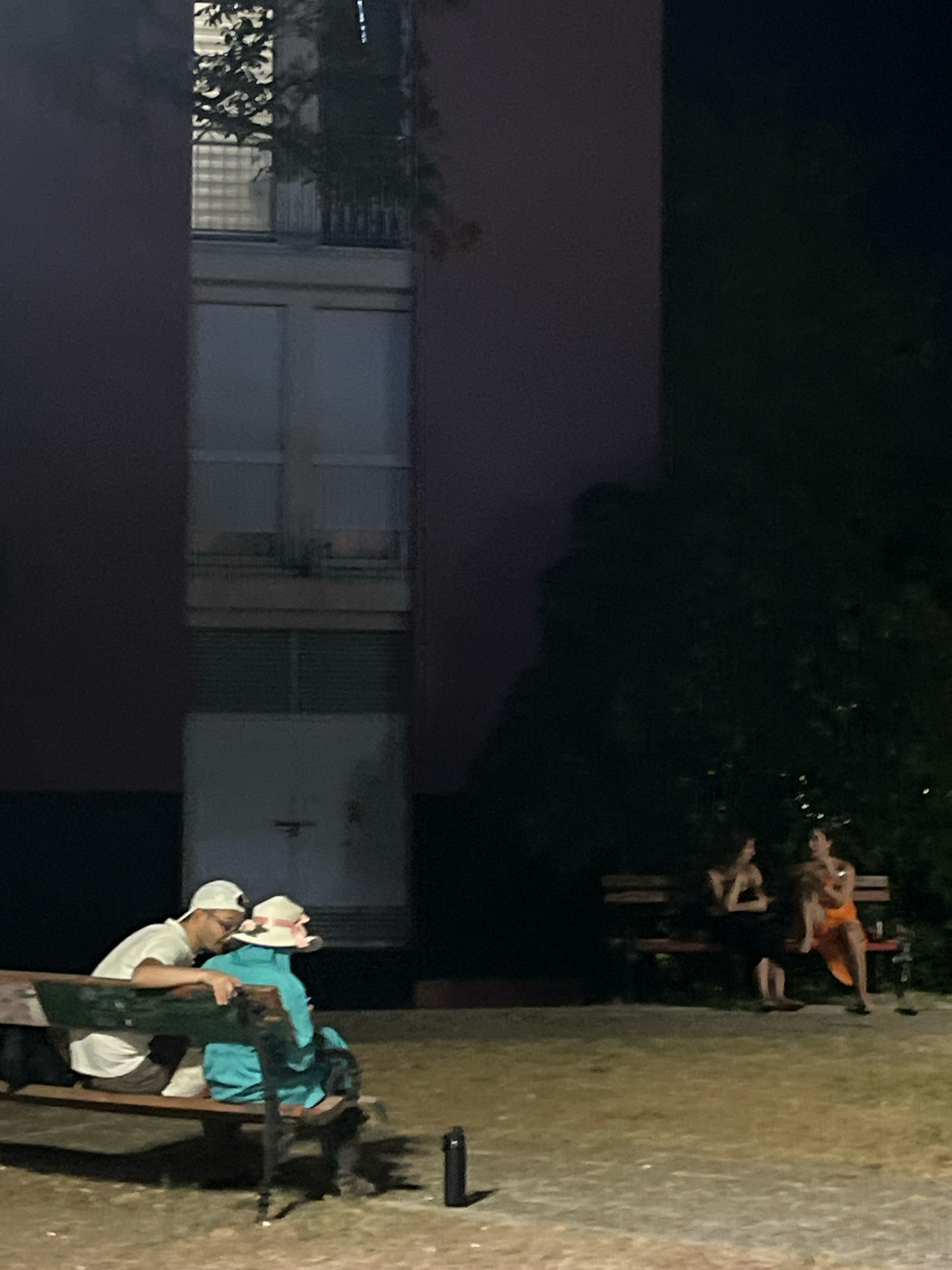 A group of people are seated on park benches in front of a dark building at night. In the foreground, on the left, a couple is seated on a wooden bench. The person on the left, wearing a white t-shirt and a white baseball cap, leans towards the person on the right. The person on the right is wearing a turquoise jacket and a wide-brimmed hat with pink ribbon. Behind them, further to the right, a group of three people are seated on another wooden bench. One person is wearing a black dress, and another is wearing an orange dress. The building behind them appears to be a multi-story structure with visible windows and balconies. The area is illuminated by artificial light, casting shadows. The ground is a mix of grass and dirt. The image is taken at night in an outdoor urban setting.