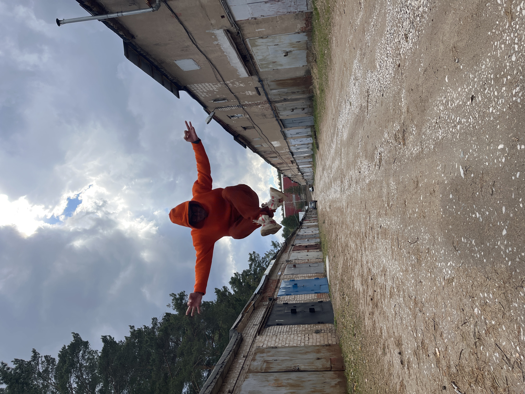 A person dressed in an orange hooded tracksuit is captured mid-air, appearing to jump or float. The person wears white sneakers with red accents. One hand is raised, forming a peace sign, while the other is extended downwards with an open palm.
The scene features a long row of utilitarian garage-like buildings or storage units, constructed from brick and concrete with various metal doors. These structures appear somewhat weathered. A dirt path, covered with small white pebbles and patches of dry grass, runs alongside the buildings. In the background, green trees are visible beneath a sky filled with grey and white clouds, with some patches of blue.
The photograph is intentionally rotated 90 degrees clockwise, presenting the ground and buildings as vertical surfaces. When viewed in its intended orientation, the person would be jumping across a dirt path in front of the garages. The activity conveys energy, playfulness, and confidence.
The time of day is daytime, with natural, diffuse light suggesting a cloudy or partly cloudy sky. The location is an outdoor setting, likely an urban or industrial periphery in Chaikovsky, Russia, characterized by older storage facilities. No visible text is present in the image.