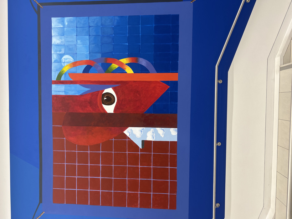 An abstract painting features geometric shapes and bold colors. A large red form with a visible eye dominates the center, positioned over a grid of red squares. Above it, a rainbow of curved lines sits over a horizontal red stripe against a background of blue squares. Below the red form, a small white arrow points towards a sky of puffy white clouds. The artwork is framed in blue, and a section of a white wall with a handrail is visible on the right side of the image.