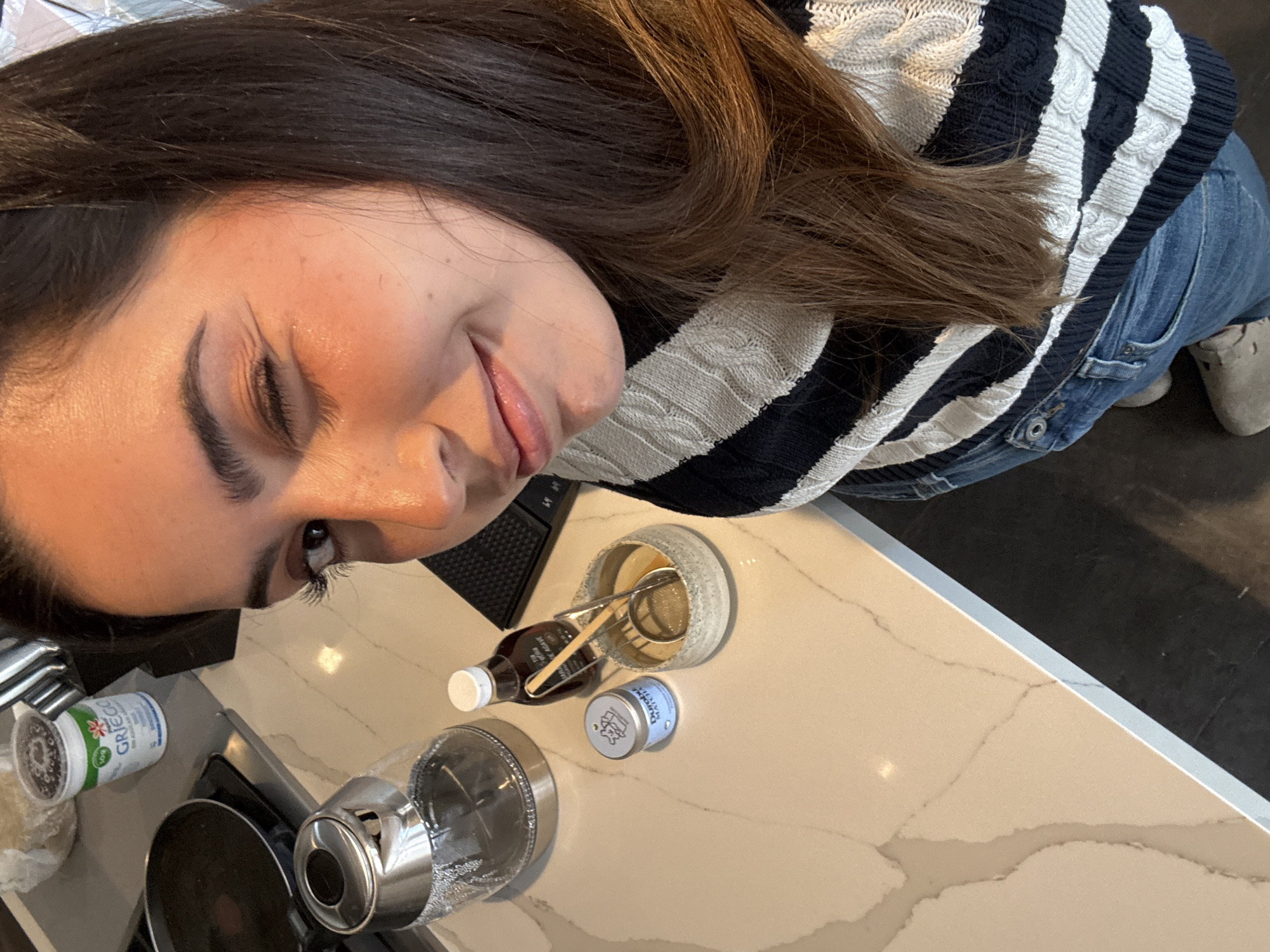 A woman is facing the camera, tilted, with dark hair and wearing a striped sweater and jeans. On a marble countertop, there is a clear glass teapot with a silver top, a green and white container, a brown bottle, a small blue container, and a cup with a strainer and spoon. There is a black object behind the cup. The location of the image is indeterminable, so the city of San Bernardino Tlaxcalancingo, Mexico cannot be confirmed or disproven.