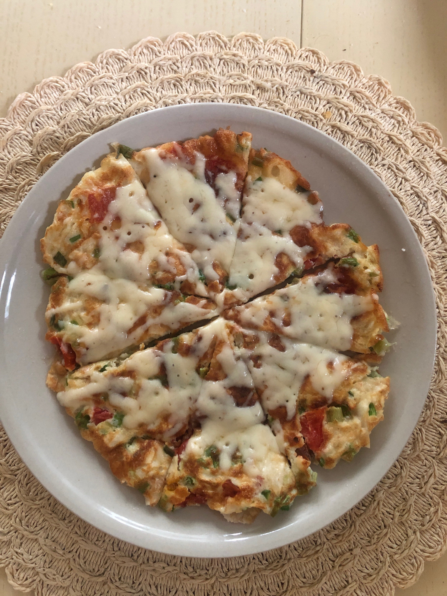 A pizza-like egg dish, sliced into eight pieces, is presented on a white plate, which rests on a beige woven placemat. The dish features a golden-brown crust with visible chunks of red tomatoes and green scallions, topped with melted white cheese. The placemat has a decorative scalloped edge and is placed on a light-colored wooden surface. The scene appears to be an indoor setting in Arfa, Russia, likely a kitchen or dining area, with natural light illuminating the food. There are no people or animals present in the image.
