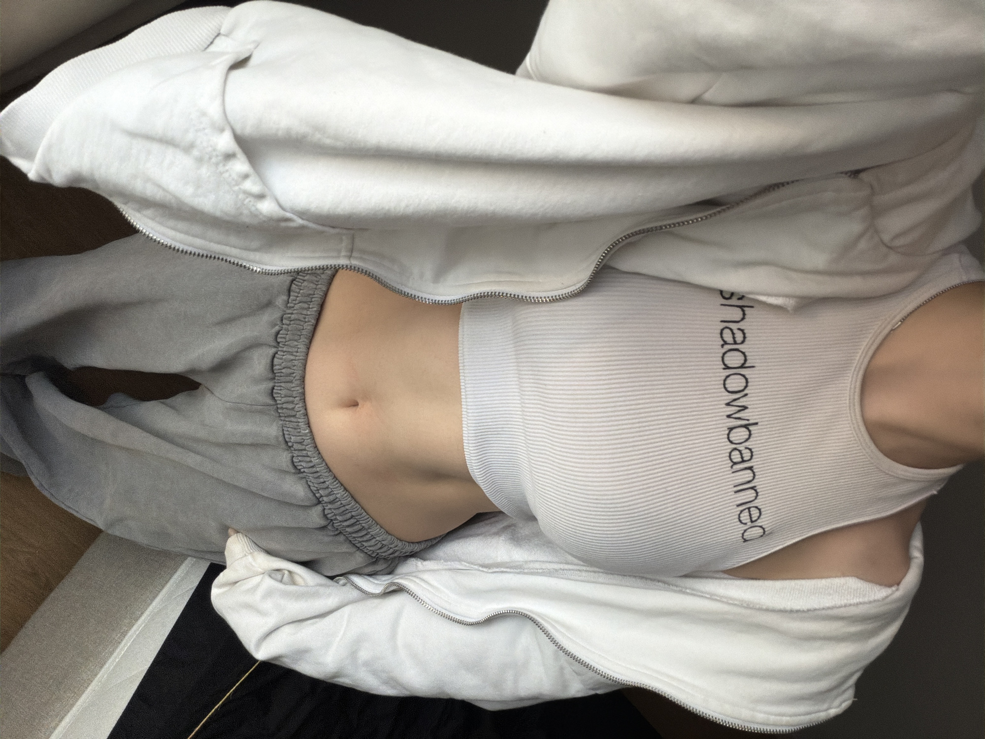 The image depicts a person lying on their back, possibly on a bed, wearing gray sweatpants and a white ribbed tank top with the text "shadowbanned" printed on it. They are also wearing a partially unzipped white hoodie. The background is slightly blurred, showing parts of a wooden floor and dark fabric. The environment does not suggest any specific location or city, and there is no information to associate this image with Moscow, Russia.