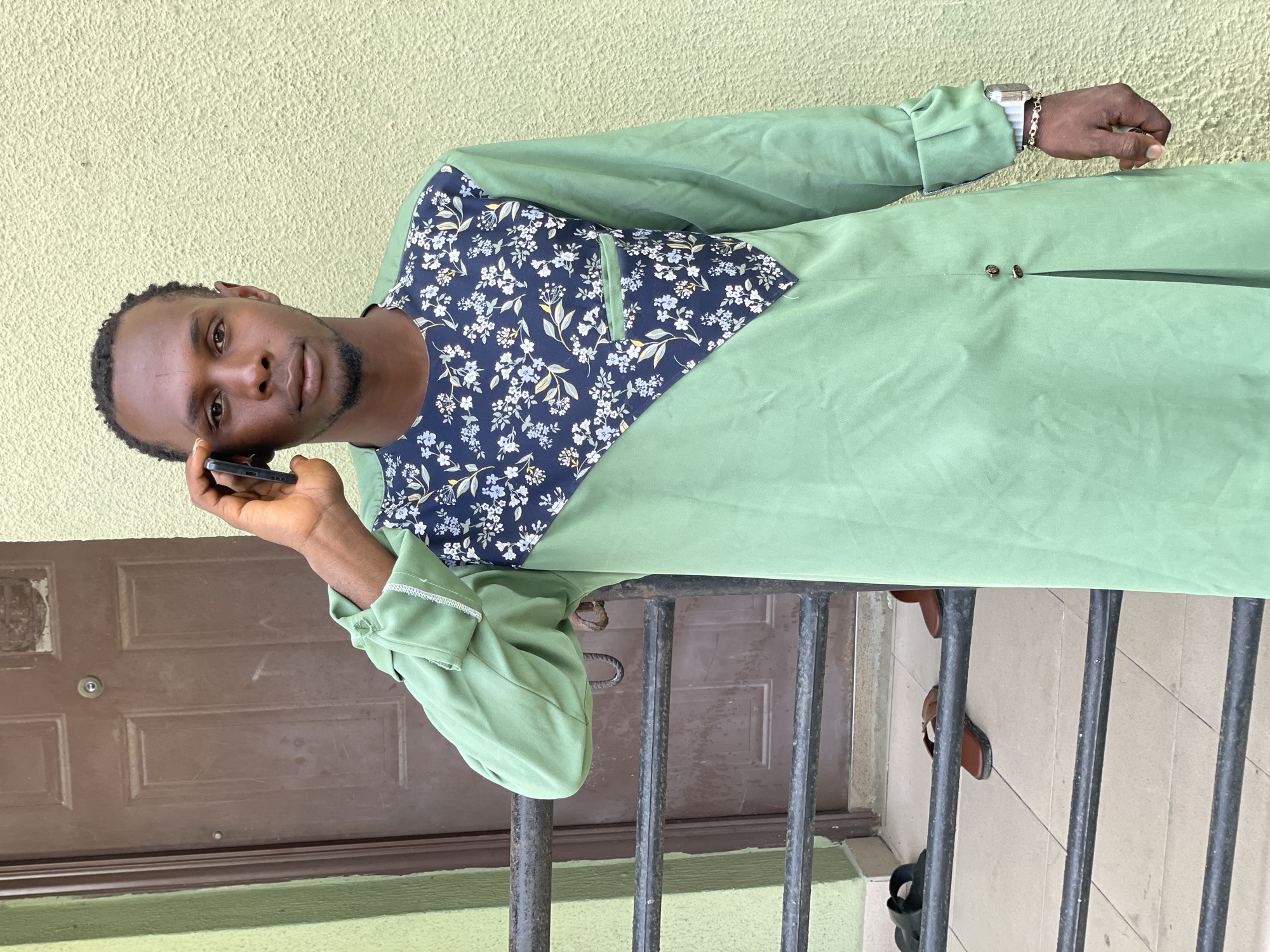 In Ado, Nigeria, a young man stands outdoors, talking on a mobile phone. He is wearing a light green tunic with a dark blue, floral-patterned chest panel. The tunic has a slit at the front and is accented with small buttons. He has a watch on his left wrist and a gold bracelet on his right wrist. He is standing in front of a brown door, with a textured, light green wall to his right. A black metal railing is in the foreground, and a pair of brown sandals and black flip-flops are visible on the tiled ground beneath it.