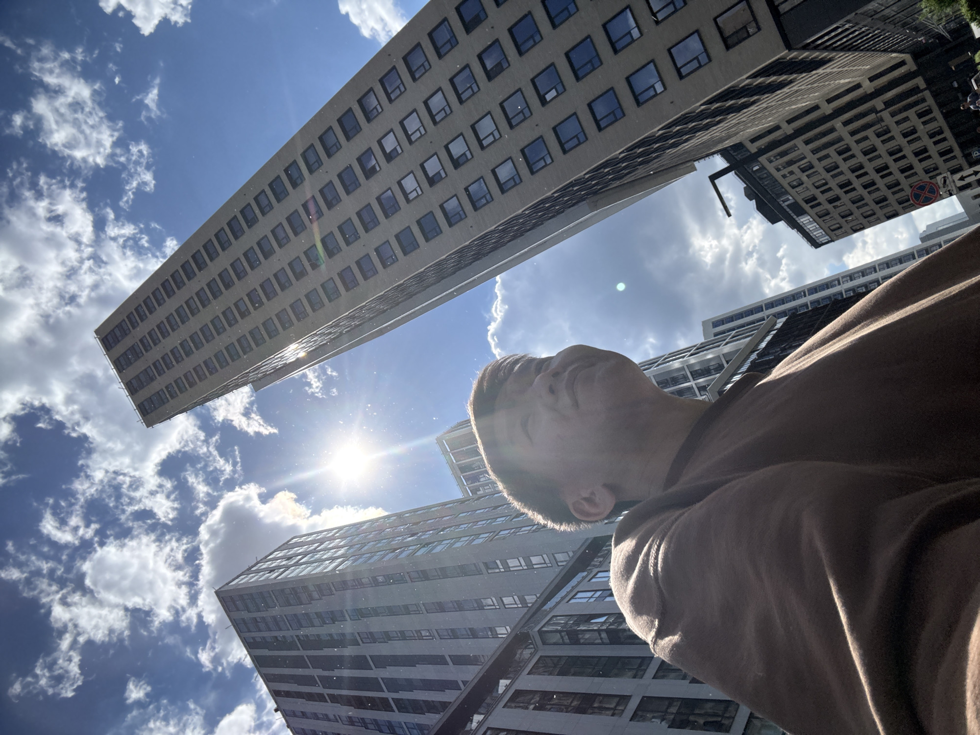An extreme low-angle shot captures an urban landscape under a bright, sunny sky. The primary subject is a person, likely male, positioned in the foreground and right side of the frame. Their head is tilted sharply upwards, looking towards the sky and the sun, with their face partially obscured by the strong backlighting. They appear to be wearing a brown hooded garment.
Towering, modern skyscrapers dominate the background and mid-ground, stretching upwards and diagonally across the frame. A particularly tall, slender building with numerous rectangular windows extends across the left side, appearing to lean into the sky due to the wide-angle perspective. Other multi-story buildings with similar architectural styles are visible in the background, their facades reflecting the bright light.
The sky is a vibrant blue with scattered white, fluffy clouds. A brilliant sun is positioned in the upper-middle left, casting intense light and creating a prominent lens flare, adding a radiant quality to the image. Small, illuminated particles, possibly dust or moisture, are visible in the air, particularly around the sun's rays. The strong sunlight and clear sky indicate a bright day, likely midday or early afternoon. The scene depicts a bustling, modern urban environment, consistent with city centers in places like Moscow, Russia. No discernible text is clearly visible in the primary image.