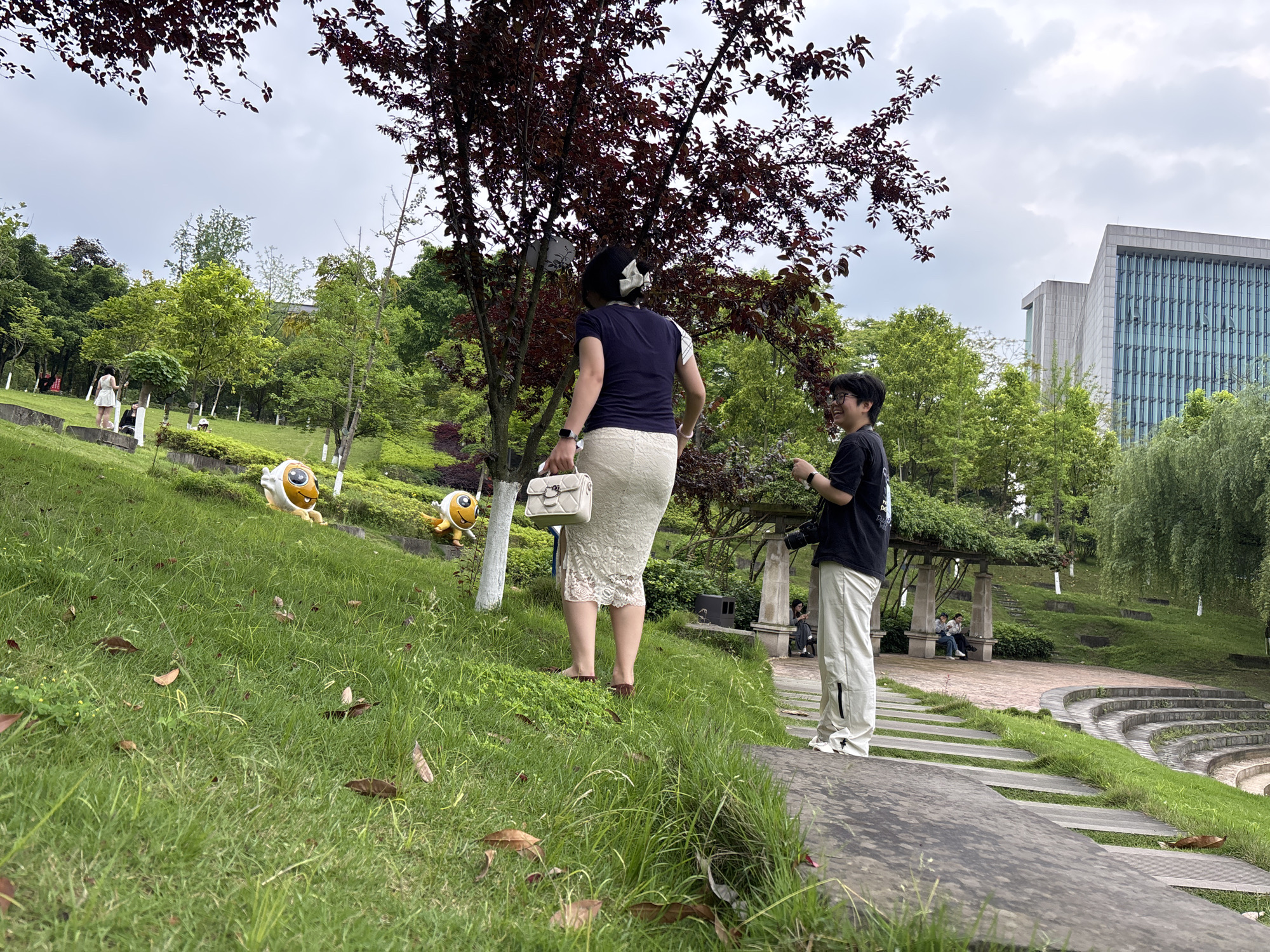 A daytime shot shows two people on a grassy hill. The person on the left is a woman wearing a dark blue short-sleeved shirt, a white lace skirt, and sandals. She carries a white purse and has a white bow in her hair. She is standing barefoot in the grass. To her right, standing on a stone platform, is another person wearing a black t-shirt, white pants, and white sneakers. This person appears to be holding a camera. Behind them is a tree with reddish-purple leaves. The background features a green grassy hill with cartoonish yellow creatures with large eyes. In the distance, there are other people in a shaded seating area and a modern building. The scene appears to be a park in Jiangjin, China.