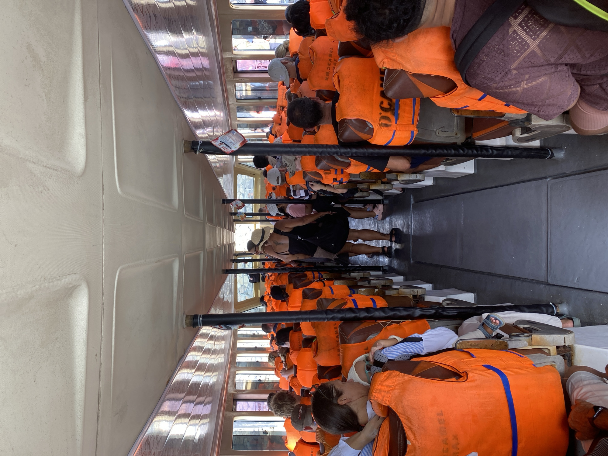 This media file captures an interior view of a crowded boat, likely a ferry or tour boat, filled with passengers wearing bright orange life vests. The perspective is from within the boat, looking down the length of the seating area. The people are seated on what appear to be bench-style seats, facing forward in rows. Many individuals are seen from the back or side, with a few faces visible, particularly in the foreground and towards the back. The overall mood seems relaxed, with some passengers resting or looking out.
The scene is set inside a vessel with windows lining the sides, suggesting a daytime journey, possibly under fair weather conditions. The interior of the boat is utilitarian, with visible structural elements and simple seating. The dominant color is the vibrant orange of the life vests, creating a strong visual contrast with the more muted tones of the boat's interior and the passengers' clothing.
Notable details include the orderly rows of life vests, indicating a safety-conscious environment. The passengers appear to be a mix of ages and ethnicities. In the foreground, a person's feet are visible, clad in sandals. Towards the back, there are glimpses of the outside through the windows, hinting at a water-based journey.
There is visible text on some of the life vests. One vest clearly shows "KAMEI MAX" in black lettering, and another has "D.C." printed in blue. The setting is identified as Denpasar, Indonesia, which aligns with a tropical or subtropical maritime environment often associated with boat travel. The overall impression is that of a group of tourists or travelers on an excursion in a warm climate.