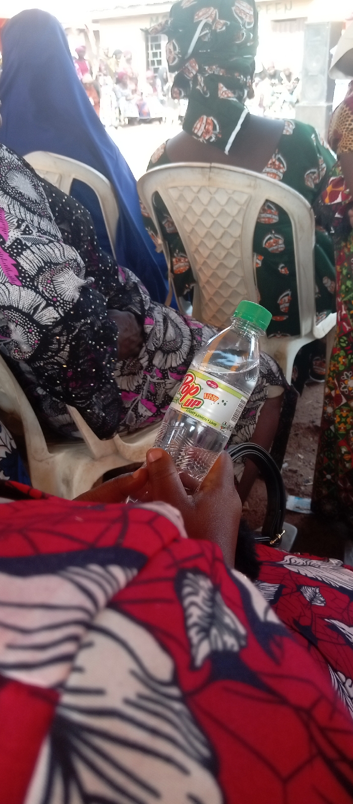 The scene is an outdoor gathering in Kasuwan Magani, Nigeria. The viewpoint is from a seated individual, looking forward and slightly to the right. In the immediate foreground, a person with dark skin holds a clear plastic bottle of "Pop Up Fizzy Light Lemon + Lime" with a green cap. This person wears a red, white, and black patterned fabric. Directly in front, another individual is seated on a white plastic chair, their back to the camera, wearing a dark green patterned dress with orange and white motifs, and a matching headwrap; a white face mask or cloth hangs around their neck. To the left of this individual, another person is partially visible, wearing a blue head covering and a black, white, and pink patterned garment, also seated on a white plastic chair. On the far right, the arm and side of a third person, dressed in multi-colored patterned fabric, are visible. The background reveals a light-colored building with a multi-paned window, and a large dark speaker is positioned to its right. Numerous blurred figures are visible in the distant background, indicating a crowd. The ground appears to be unpaved dirt.