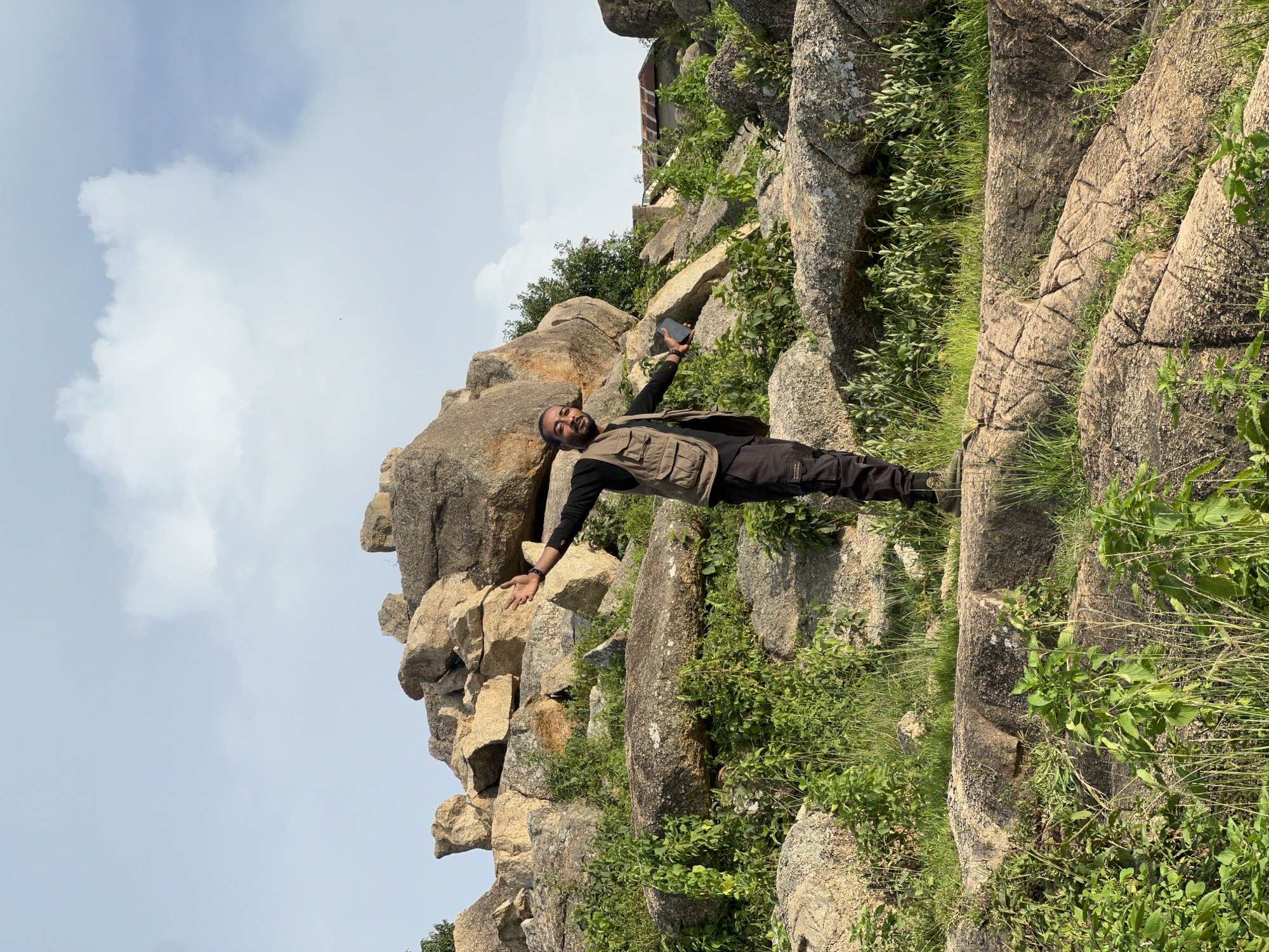 A person stands with arms outstretched on a rocky incline covered in sparse green vegetation. The individual is wearing a dark long-sleeved shirt, a light brown utility vest, and dark pants. They are holding a dark rectangular object, possibly a phone, in their right hand. The background consists of large, weathered rock formations and a partly cloudy sky. The scene is outdoors and appears to be a natural, undeveloped area, consistent with locations found on the outskirts or within natural reserves in cities like Jos, Nigeria, known for its hilly terrain and rock formations.