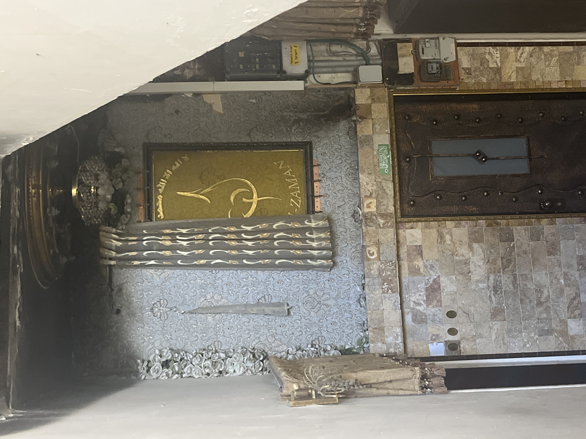 The image shows an indoor space. A golden-framed wall decoration with Arabic script is visible, along with other decorative elements, including floral motifs and wave-patterned designs. To the right, there is a metallic door or panel with a simple rectangular window, set within a wall tiled with square stones. Electrical boxes and conduits are visible near the ceiling. The lower portion of the frame contains what looks like a stack of frayed linens. The location, Tel Aviv, Israel, is only suggested by the image title and is not directly verifiable from the visible content.