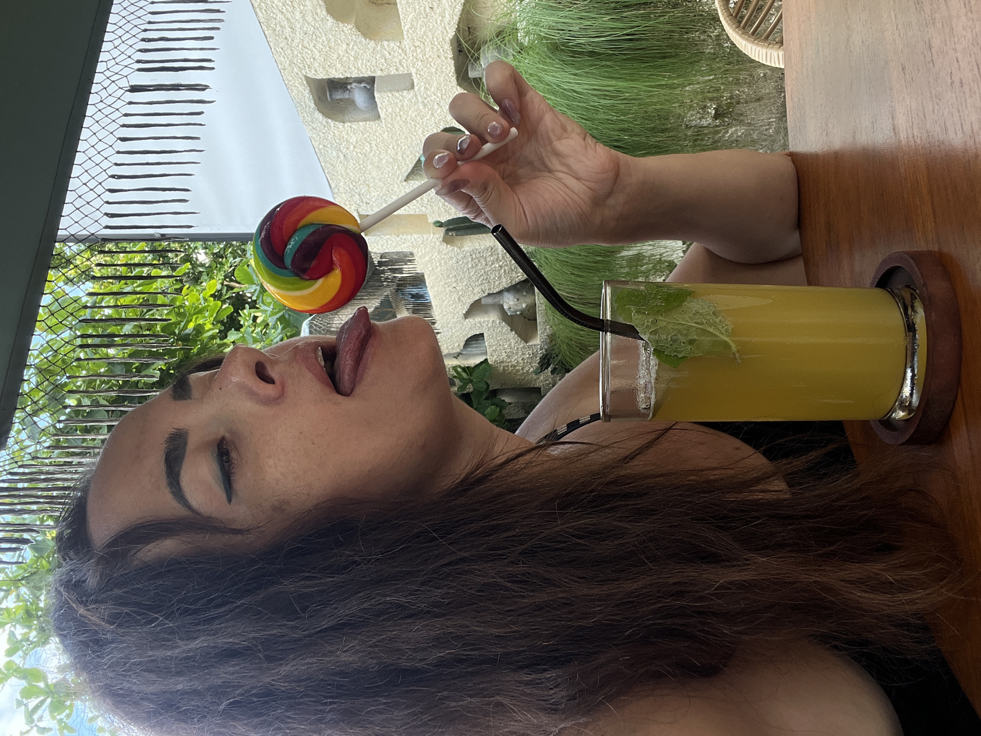 A woman with dark hair licks a rainbow swirl lollipop while holding it with her right hand. She is seated at a wooden table with a glass of yellow liquid and mint leaves. She is wearing a black top. The background shows greenery and a white textured wall with arched cutouts. The scene is outdoors, with sunlight visible. The overall setting appears to be a cafe or restaurant in Kuta Selatan, Indonesia, characterized by tropical foliage and the distinctive architecture.