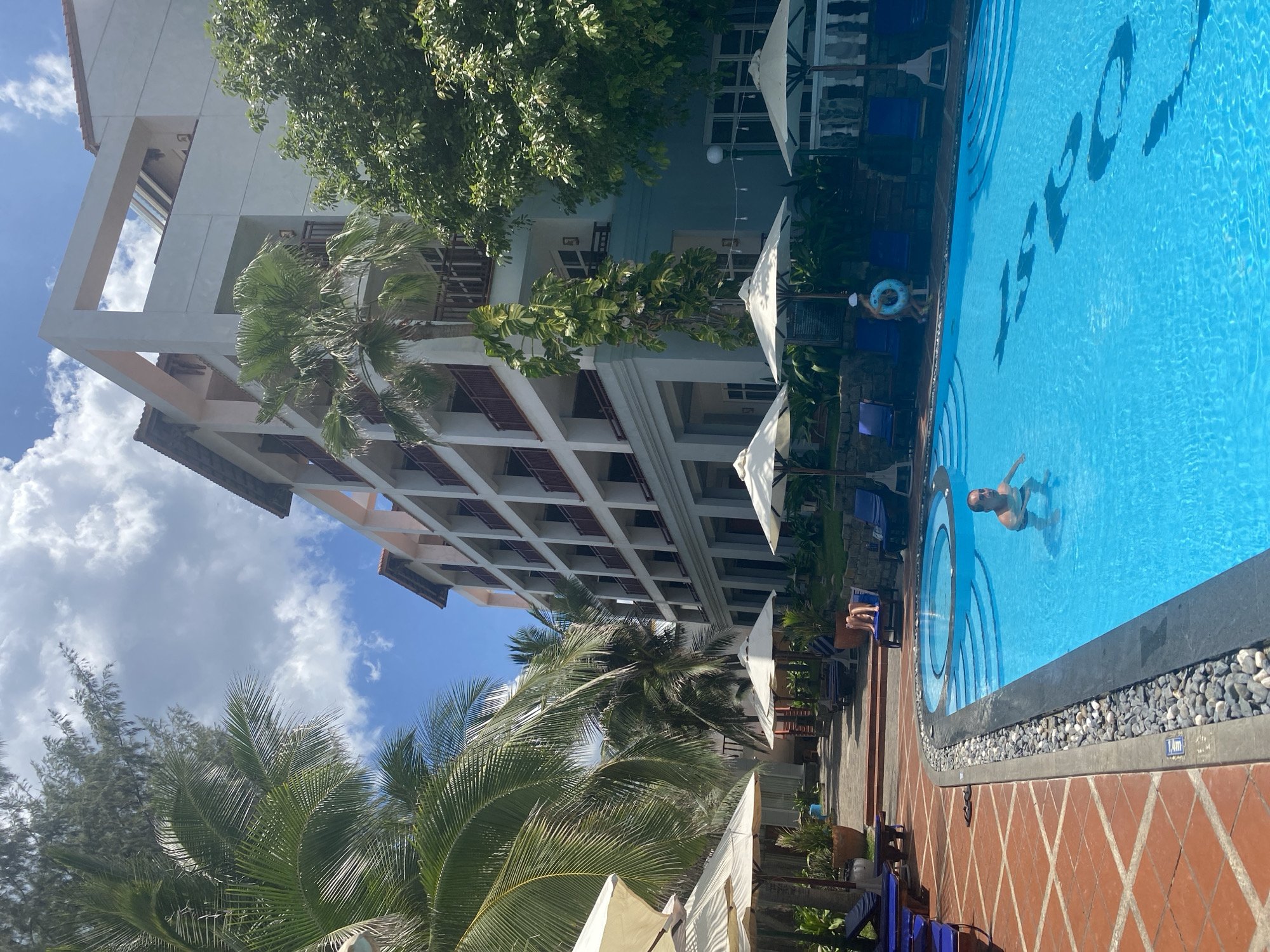 The image captures a bright, sunny day at a resort in Phu Thuy Ward, Vietnam. The scene is dominated by a large, clear blue swimming pool, with the depth markings "1.4m" visible on the edge. A man is swimming in the pool, appearing relaxed. In the pool, the numbers "15-20" are visible as part of the pool's design, likely indicating a depth range or a section of the pool.
Adjacent to the pool, a multi-story building with a white facade and multiple balconies with wooden shutters suggests it is a hotel or resort. Palm trees and lush green foliage surround the building and pool area, adding to the tropical atmosphere. Several white umbrellas are set up around the pool, providing shade. Blue lounge chairs are also visible, with one person relaxing on a chair by the pool. The ground around the pool is paved with terracotta tiles arranged in a diamond pattern. The sky is blue with scattered white clouds, indicating fair weather. The overall impression is one of a leisurely vacation day in a tropical environment.