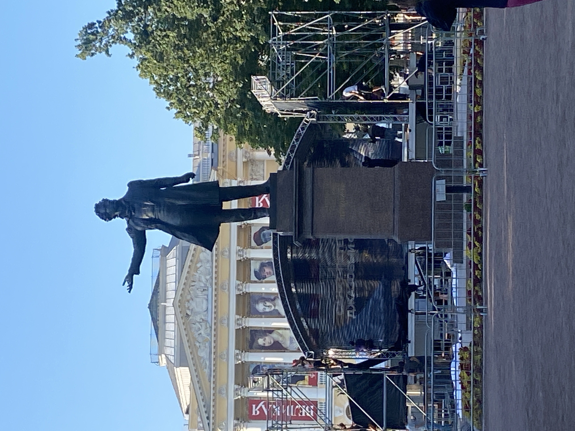 The image captures a full-body bronze statue of a man dressed in a formal coat and trousers, standing on a stone pedestal. The statue is positioned outdoors in front of a classical-style building with columns and a triangular pediment. The building appears to be a museum or historical landmark, possibly the Russian Museum, given the presence of large banners with portraits of individuals on its facade.
In the foreground, a temporary stage structure is being assembled or dismantled. Scaffolding, black tarps, and metal barriers are visible, indicating some event or exhibition is in progress or has recently concluded. Several individuals are present around the stage area, engaged in various tasks, some climbing the scaffolding and others working on the ground.
The overall environment suggests a public space in Saint Petersburg, Russia, likely a city square or courtyard. The clear blue sky and lush green trees in the background indicate it is daytime. The scene is captured from a slightly low angle, looking up towards the statue and the building. The ground in the immediate foreground is a paved area, possibly a plaza or sidewalk. Flowerbeds with red and yellow flowers are visible along the barriers.