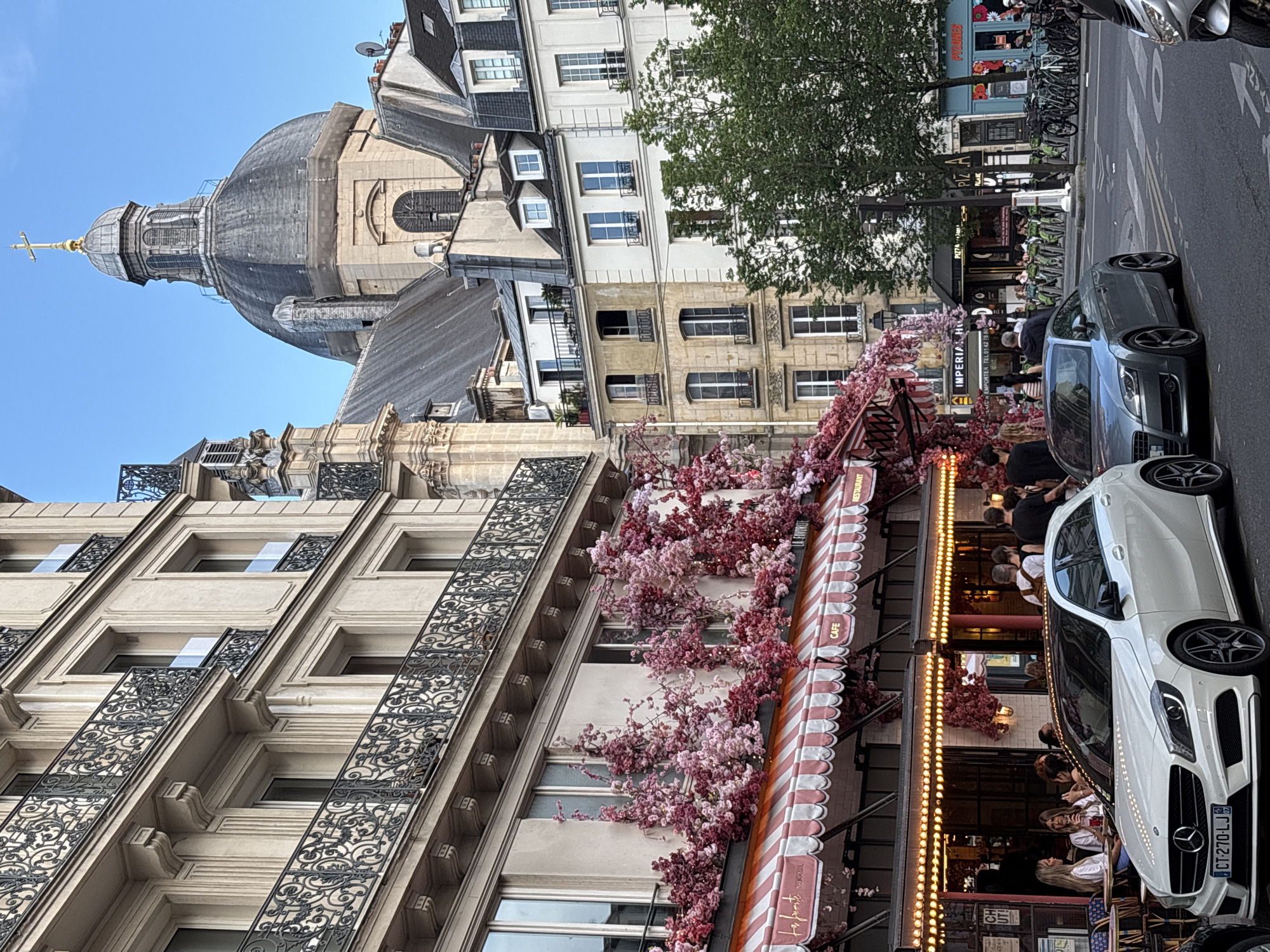 The image depicts a street scene in Paris, France, likely in a charming, historic district. In the foreground, two cars are parked on the street: a white Mercedes-Benz A-Class on the left and a grey Audi TT on the right. Behind the cars, a building with a pink and white striped awning stretches across the frame. This awning belongs to a cafe or restaurant, indicated by the words "CAFE" and "RESTAURANT" visible. The facade of this establishment is decorated with an abundance of pink and red artificial flowers, creating a vibrant display.
Several people are seated at tables outside the cafe, appearing to enjoy drinks and conversation. Others are walking past, suggesting a bustling atmosphere. The building behind the cafe is tall and ornate, with balconies featuring intricate wrought-iron railings. Further in the background, the prominent dome of a church, topped with a golden cross, rises above the Parisian rooftops. The sky is a clear blue. To the right, more buildings and trees line the street, and several bicycles are parked, hinting at a mode of transportation common in the city. The overall scene conveys a lively and picturesque Parisian ambiance.