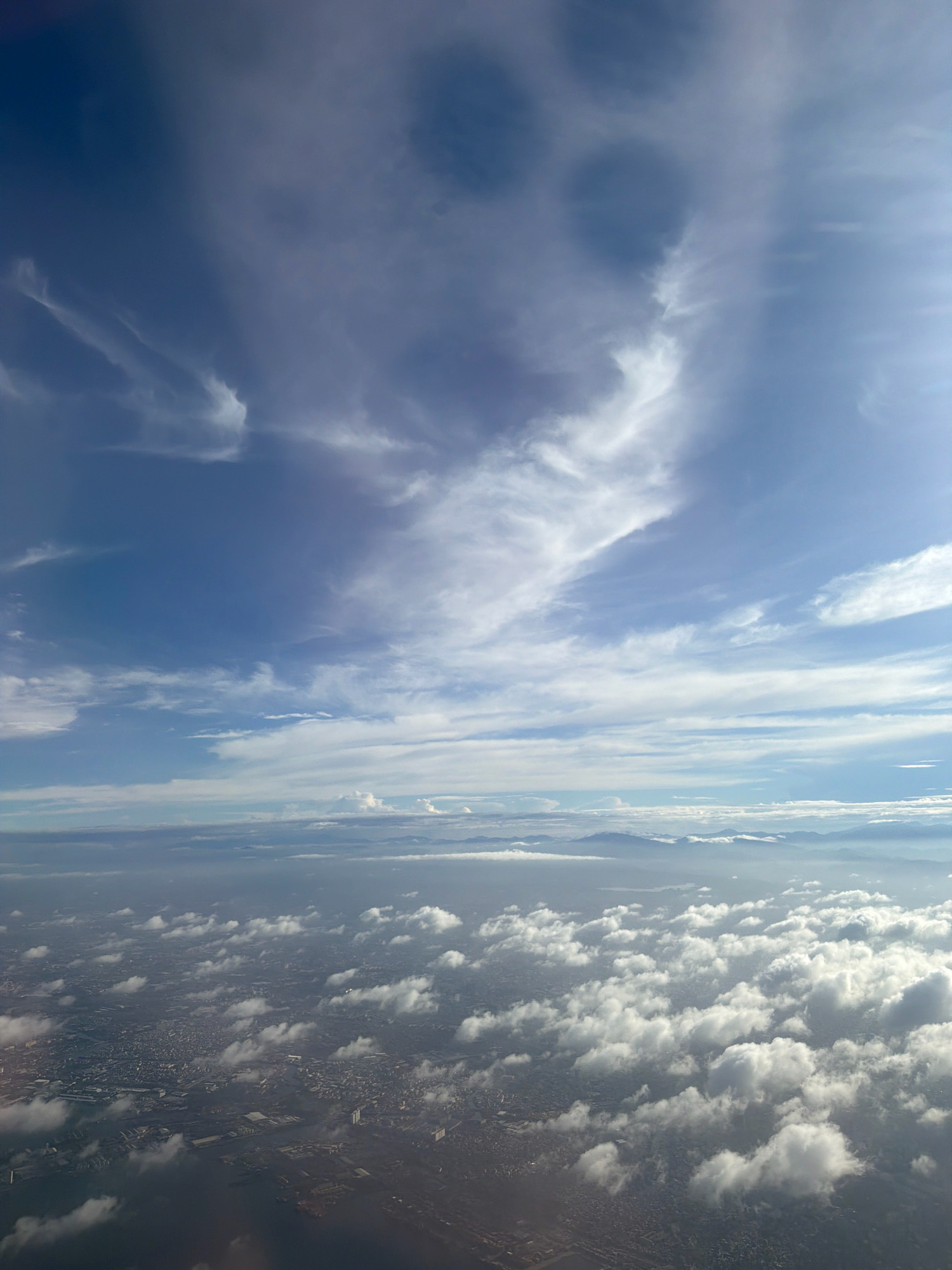 An aerial view captures a sprawling urban landscape of Manila, Philippines, beneath a dramatic sky filled with wispy clouds. The city, densely packed with buildings, stretches towards a hazy horizon where mountains are faintly visible. Patches of fluffy white clouds drift over the urban expanse, casting shadows and adding depth to the scene. The overall impression is one of a vast, busy city viewed from high above, with the sky providing a dynamic backdrop.