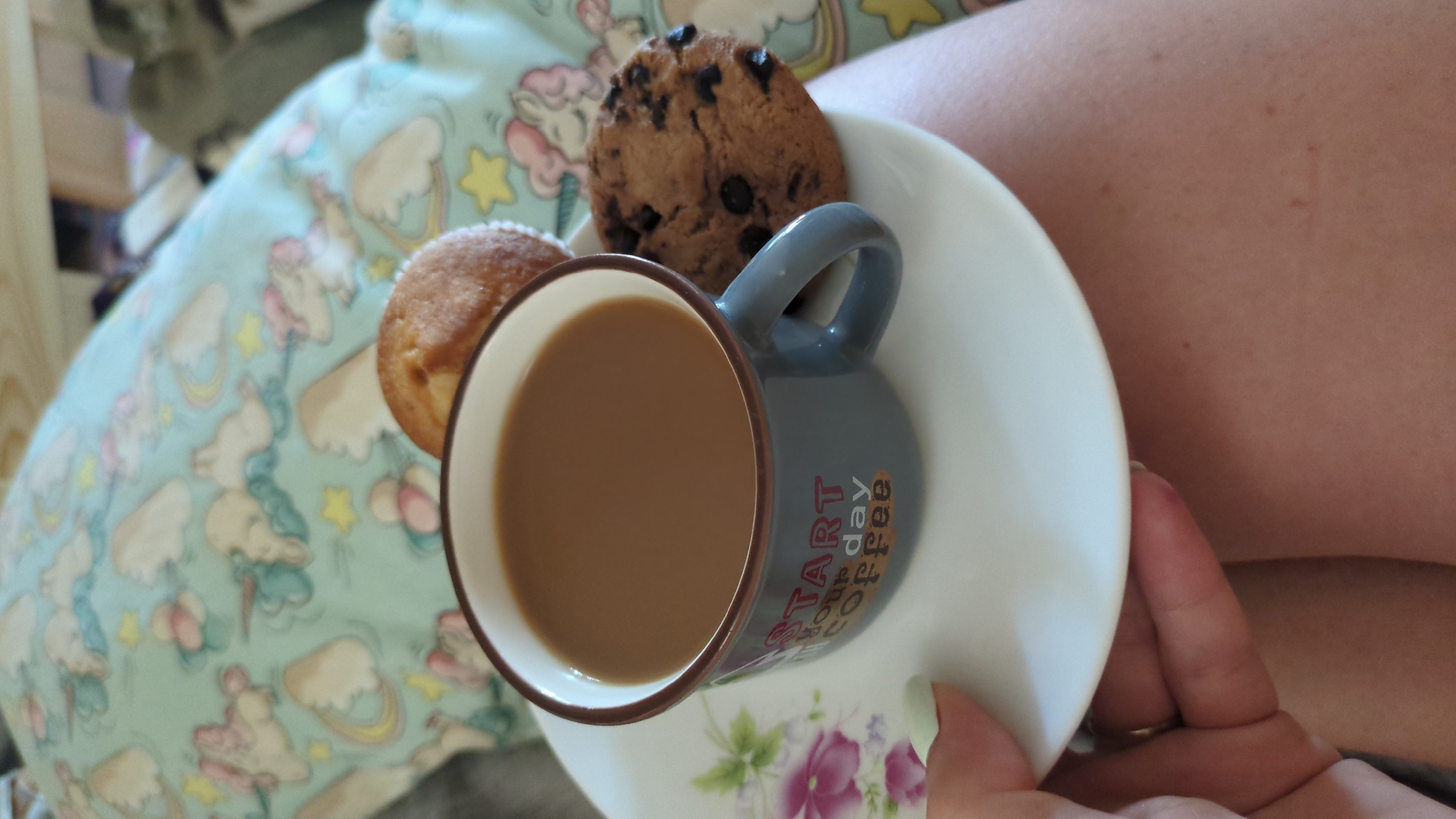 A close-up shot features a gray mug with the text "START YOUR DAY COFFEE" printed on it, filled with a brown beverage, presumably coffee. The mug is on a white saucer with a floral design. Two baked goods are placed on the saucer next to the mug: a small muffin and a chocolate chip cookie. The background consists of a pillow with a pattern of unicorns and clouds. A person's hand with light green nail polish is visible in the lower left corner, holding the saucer. The image appears to be taken indoors, possibly in a bedroom or living area. The lighting is natural. There is no discernible information in the image that would allow for the determination of its location within Chișinău, Moldova.