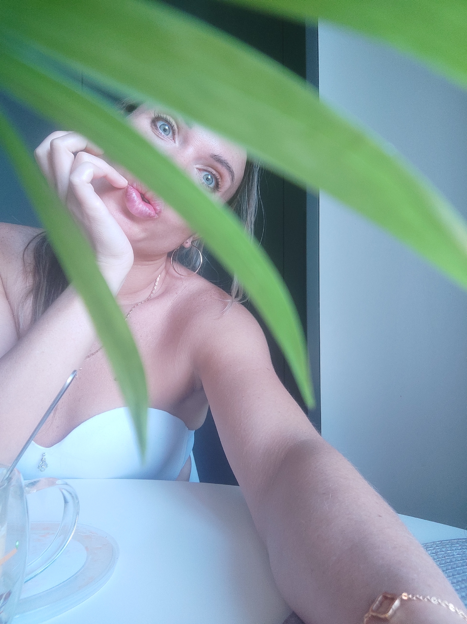 A woman is seated at a white table, with a clear glass cup and saucer visible in the lower left corner. She is positioned with her arm extended towards the camera, and her face is partially obscured by green foliage. The woman has fair skin, blonde hair, and blue eyes. She is wearing hoop earrings and a necklace. A doorway is visible in the background. The location of the scene is not discernible in the image to confirm it being Votkinsk, Russia.