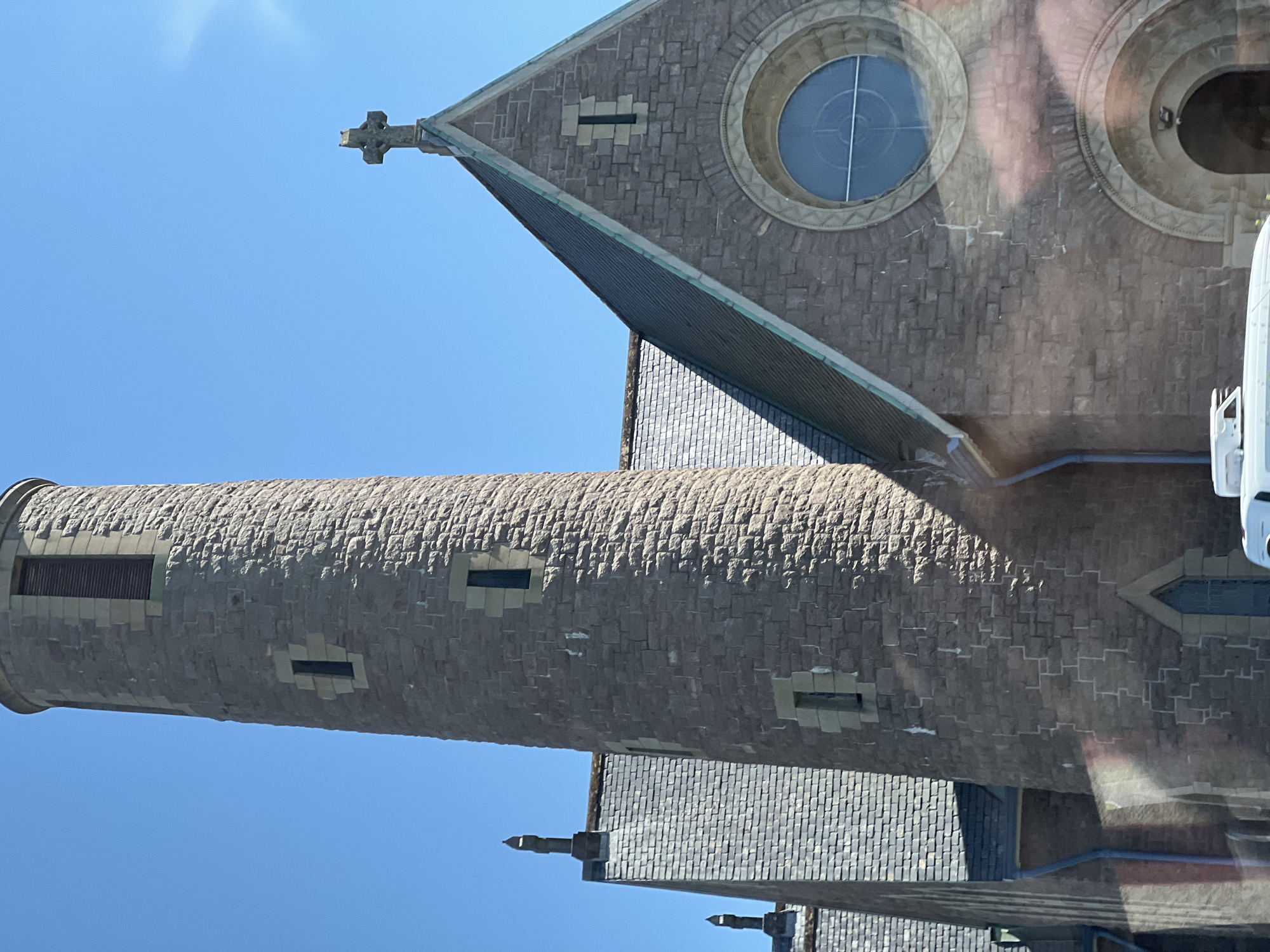 The image shows a close-up view of a stone church, possibly Saint Patrick's Church, in the city of Donegal, Ireland. The church features a tall, circular stone tower with narrow rectangular windows and a louvered opening near the top. Adjacent to the tower is the main body of the church, with a pitched, dark grey roof. The facade of the church is constructed of rough-hewn brown stone blocks, with a prominent circular window with a light blue pane and a decorative stone surround. Above this, on the gable end, is a smaller, narrow rectangular window. A Celtic-style cross is mounted at the peak of the gable. The sky is clear and bright blue, suggesting a sunny day. There are reflections on the lower part of the image, indicating it may have been taken through a window. A sliver of a white vehicle is visible at the bottom of the frame.
