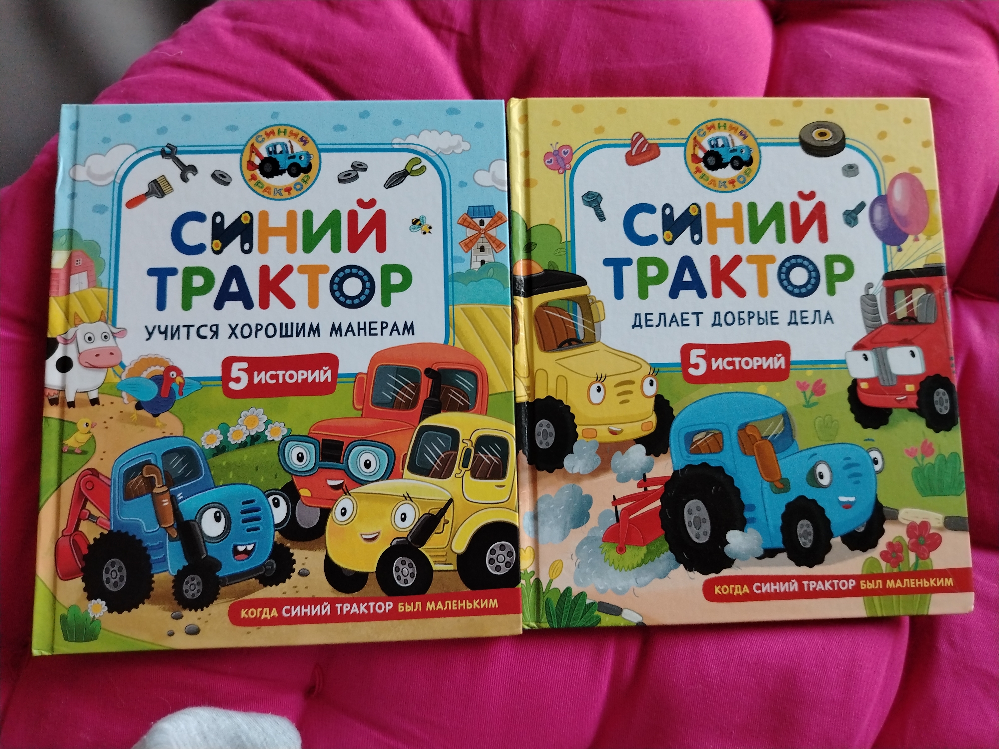 The image shows two children's books with cartoon tractors on their covers, placed side-by-side on a bright pink quilted surface. The book on the left has a blue cover with the title "СИНИЙ ТРАКТОР" (Blue Tractor) written in large, colorful letters. It features a blue tractor with a bucket loader, a red tractor, a yellow tractor, a cow, a turkey, and a duckling. The text below the title reads "УЧИТСЯ ХОРОШИМ МАНЕРАМ" (Learning Good Manners) and "5 ИСТОРИЙ" (5 Stories). The book on the right has a yellow cover with the same title "СИНИЙ ТРАКТОР". It depicts a yellow tractor, a red truck, and a blue tractor with a plow. The text reads "ДЕЛЯЕТ ДОБРЫЕ ДЕЛА" (Does Good Deeds) and "5 ИСТОРИЙ" (5 Stories). Both books have a circular emblem with a tractor inside at the top. The background is a soft, bright pink quilted fabric. The image is a close-up of the two books, and there is no indication of their location in Dubai, United Arab Emirates.