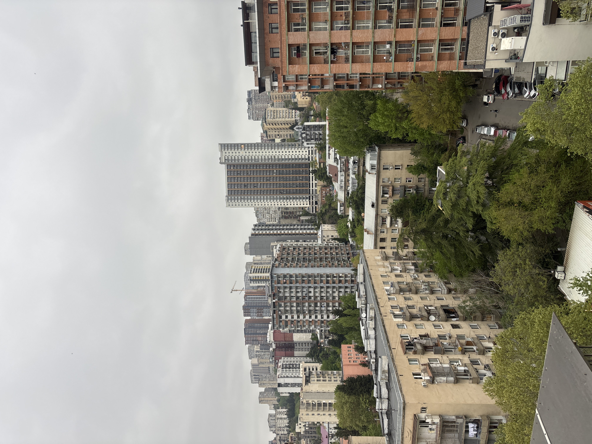 The image shows a cityscape in Tbilisi, Georgia, under an overcast sky. Tall apartment buildings of varying heights and architectural styles dominate the view, with several high-rise buildings in the midground and denser, shorter buildings in the foreground. Green trees are interspersed among the buildings and line a street below. Cars are parked on the street. The buildings exhibit diverse construction materials and colors, from red brick to concrete with windows and balconies. A construction crane is visible on the horizon among the buildings.