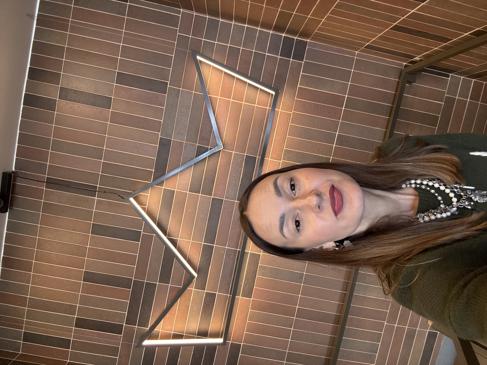 A woman with long brown hair is facing the camera. She is wearing a dark green top and a pearl and metal necklace. She is standing in front of a wall with brown brick-like tiles. There is a geometric metal light fixture on the wall. There is a partial view of a dark metal grid or shelf structure in the upper right. The location is unknown and there is no indication that the location is Sochi, Russia.