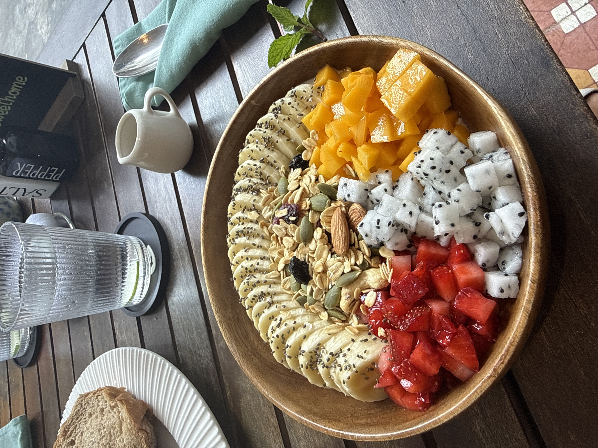 A top-down, close-up shot shows a wooden table with a bowl of fruit and other breakfast items. The bowl is filled with sliced bananas, mango chunks, diced dragon fruit, diced strawberries, and granola. The bananas are sprinkled with chia seeds.
To the left of the bowl is a small white creamer, a blue napkin, and a spoon. Below the bowl, on the left, is a glass of water, a white plate, and a slice of bread. Next to the glass of water are salt and pepper shakers, with a box that reads "sweet home" on top of them. A mint sprig is placed on the right side of the fruit bowl.
The table has a light-colored wooden plank surface. The scene suggests a casual breakfast setting, likely in a restaurant or cafe. Though the context suggests Đà Nẵng, Vietnam, there is no confirmation within the image itself.