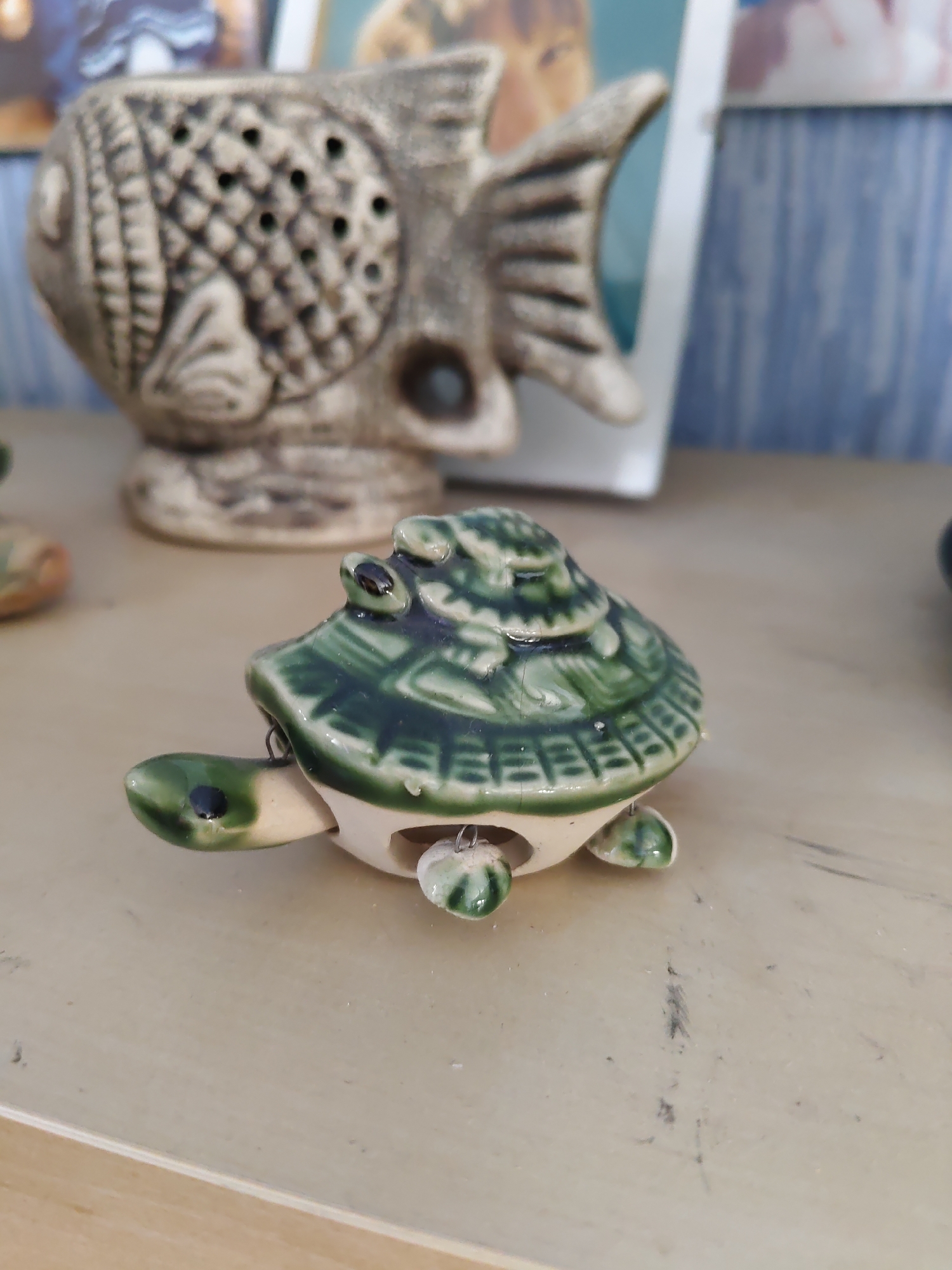 An overhead, slightly right, medium shot shows a ceramic turtle figurine positioned in the foreground on a light-colored, wooden surface. The turtle is primarily a light beige color, with its shell decorated with a glossy green glaze in a layered, patterned design. The shell has raised, segmented sections, and the green glaze has a slight crackle effect in places. The turtle's head is green with a black eye, and its neck is beige. It has four small, green feet, also with a glossy finish. A thin, silver wire loop is attached to the top of the shell, and another is visible under the turtle's neck.
In the background, slightly out of focus, is a larger, beige ceramic fish figurine with intricate detailing, including rows of small holes on its body and a fan-like tail. Behind the fish, a blurred white frame contains images, one with a hint of a person's face and the other with vertical blue and white stripes, suggestive of a patterned background. The overall environment appears to be indoors, likely a shelf or tabletop.