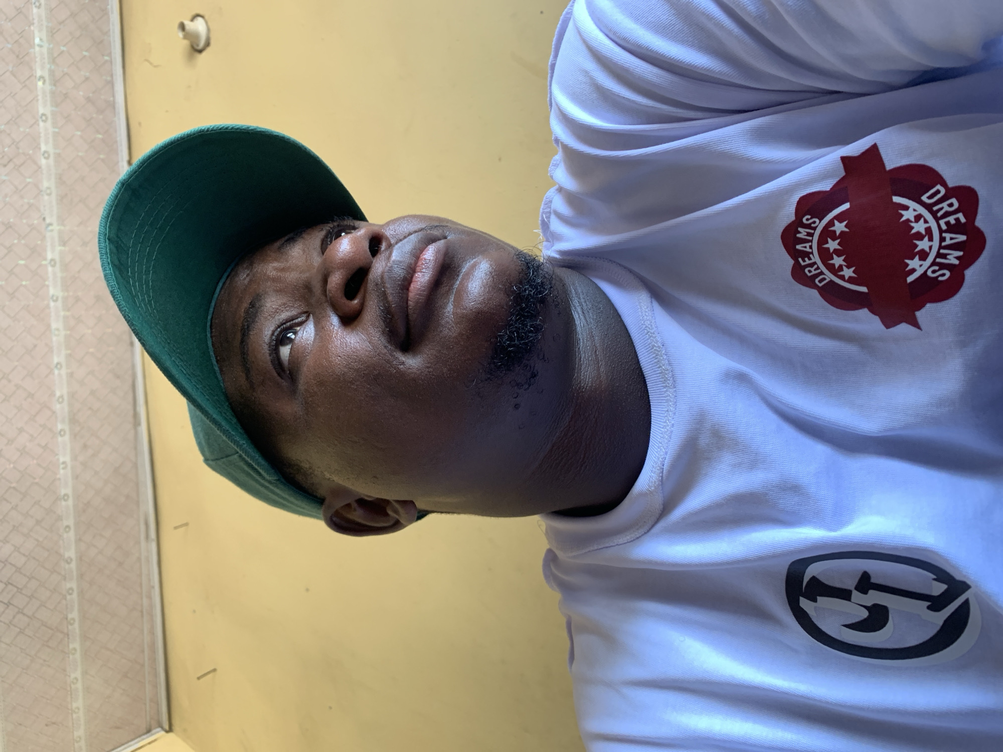 A dark-skinned man is captured in a close-up, low-angle portrait, looking upwards and to his right, with a neutral to contemplative expression. He wears a teal green baseball cap, turned backwards or to the side, and a white t-shirt. His face features a short goatee.
The white t-shirt displays two distinct logos. On the right chest area (viewer's left), there is a circular, red-burgundy emblem with a banner motif. The word "DREAMS" is repeated twice around the circle, separated by star symbols. On the left chest area (viewer's right), a circular black and white logo features an abstract design, possibly stylized letters or symbols.
The background consists primarily of a plain, light yellow or beige wall. A white, possibly wall-mounted fixture or sensor is visible on this wall. To the left of the man, a tiled surface or wall with vertical divisions can be seen, partially obscured. The lighting is bright, suggesting daytime. The man appears to be posing for a photograph. The setting is in Akwanga, Nigeria.