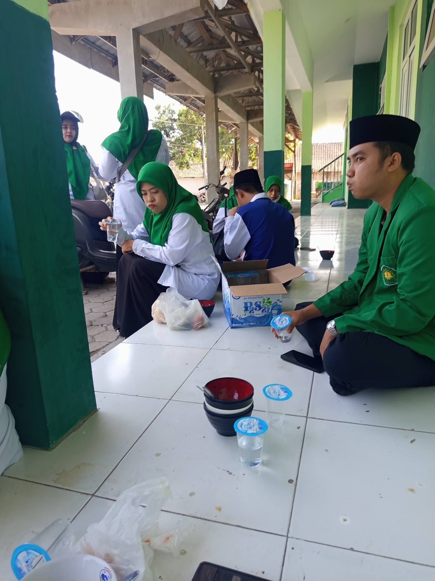 In Lumutan, Indonesia, a group of young adults, likely students based on their attire, are gathered on the tiled floor of an outdoor covered walkway, possibly on a school or university campus. The architecture features green and white painted columns and a roof structure suggesting an open-air environment.
In the foreground, a young man wearing a black peci and a green jacket is seated on the floor, looking to his right. In front of him, a small blue-lidded cup of water sits on the tile. To his left, another similar cup is empty, and a stack of bowls, one red and black, is also on the floor. A partially open cardboard box labeled "PS3 21" and containing what appear to be small plastic bottles of water is visible to the left of the young man.
Further back and to the left, a young woman in a green hijab and white shirt is seated, holding a glass of water in her right hand and looking down. Next to her are two plastic bags, one containing what looks like food items. To her left, another young woman in a green hijab and white shirt is partially obscured, seated behind her. Behind them, a motorcycle is parked.
In the background, other individuals in similar green and white attire are visible, and the scene suggests a casual gathering or break time. The overall impression is one of a relaxed, communal activity in an outdoor educational setting.